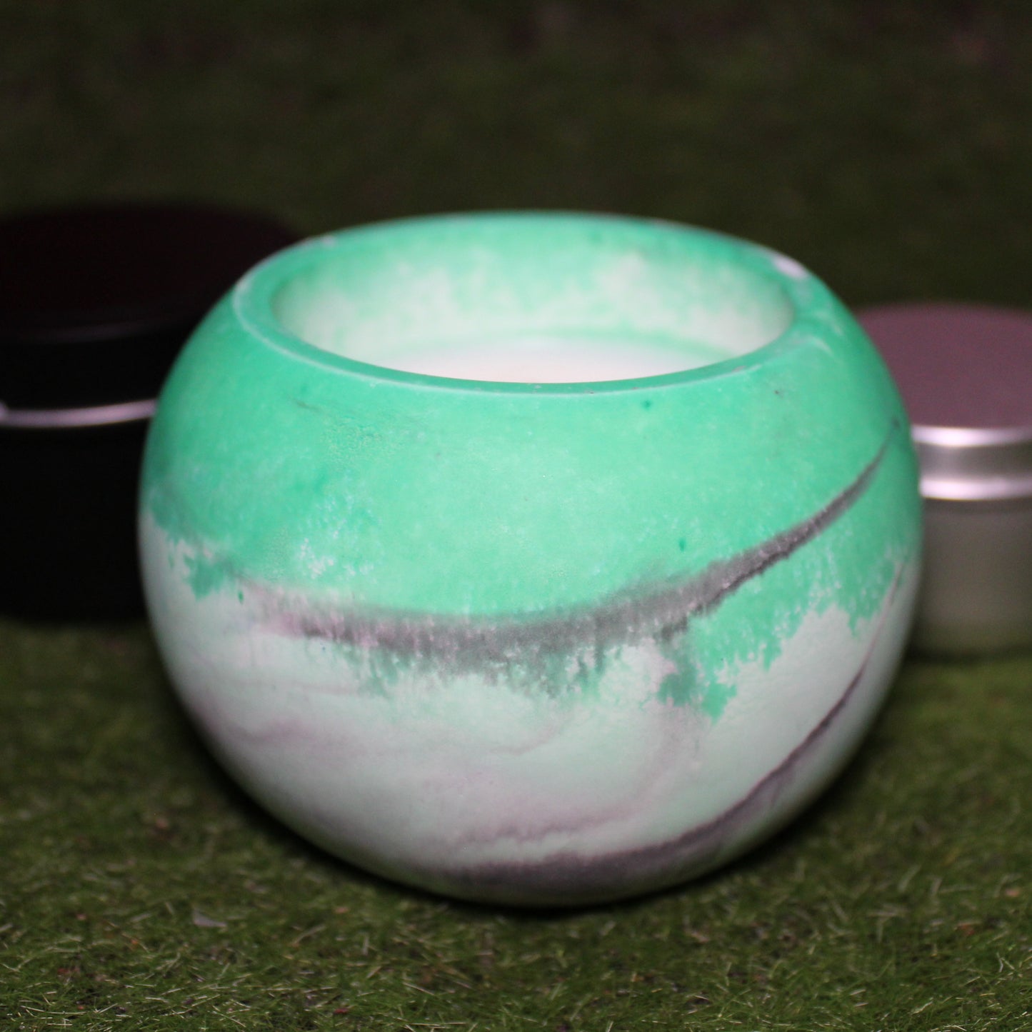 Handcrafted Ecocrete candle holder featuring the black, white, and green colors of the agender pride flag blended smoothly together. Product shot on grass with styling that includes a silver 2oz candle tin and black 4oz candle tin behind it, displaying the three available sizes of this LGBTQIA+ home decor piece.