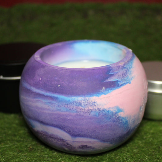 Handcrafted Ecocrete candle holder featuring the pink, blue and purple colors of the bisexual pride flag blended smoothly together. Product shot on grass with styling that includes a silver 2oz candle tin and black 4oz candle tin behind it, displaying the three available sizes of this LGBTQIA+ home decor piece.