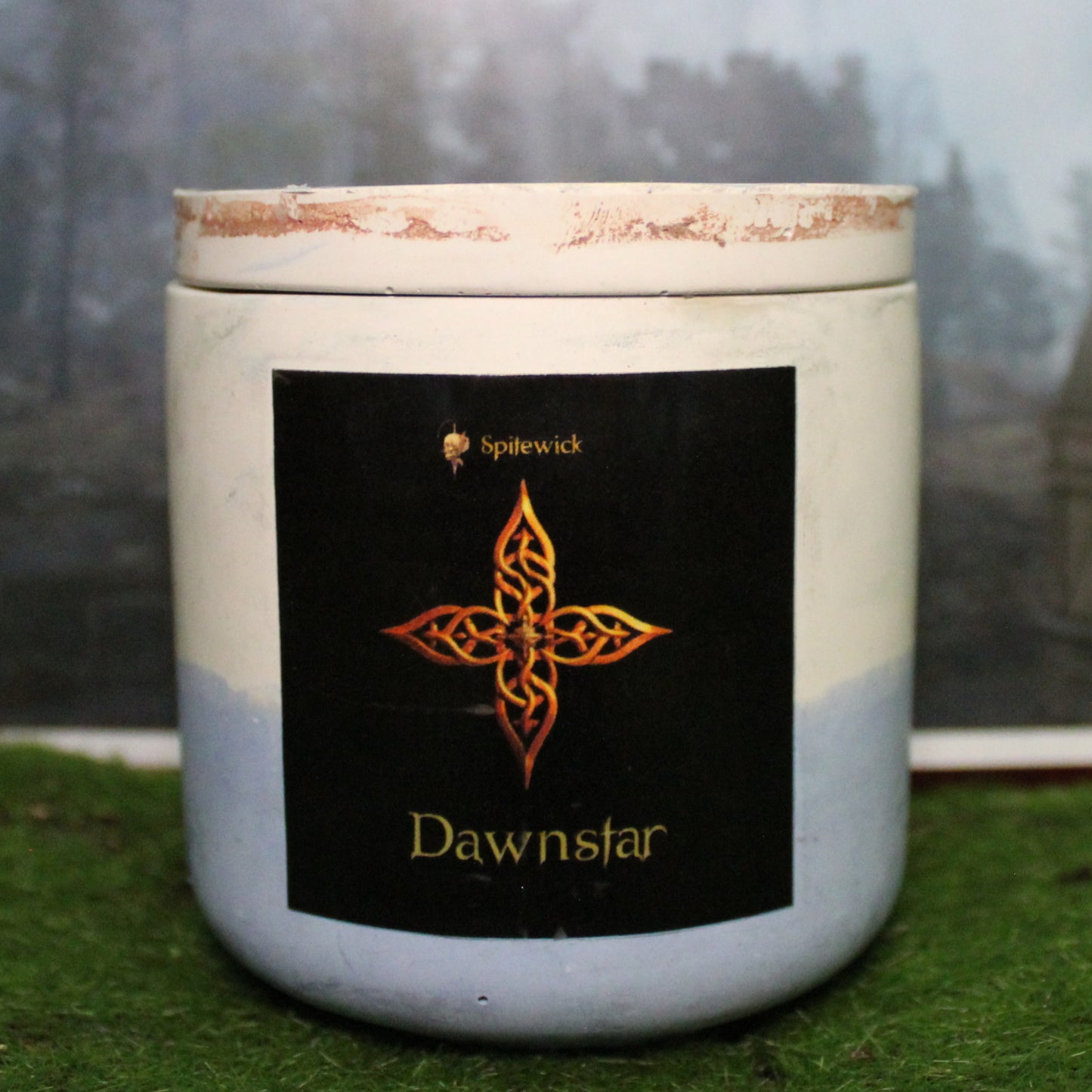 Front view of a handcrafted white and pale blue candle holder with a black label featuring a four pointed star in gold Celtic knotwork. The name dawnstar is elegantly printed in gold beneath the design. Set on grass, this piece blends artisan craftsmanship with bold identity, making it perfect for unique home decor, gifts, and statement lighting.
