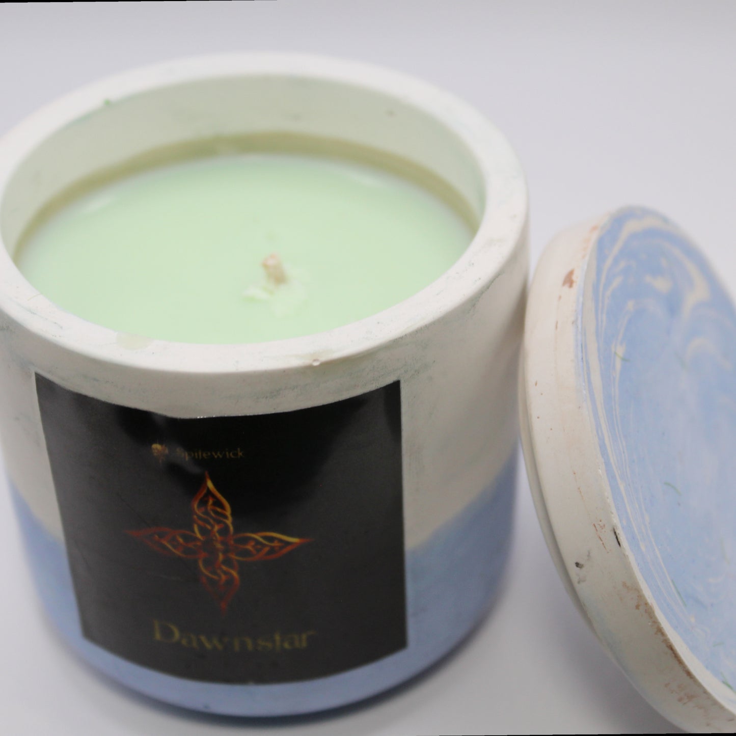 3/4 view of a white and blue handcrafted candle holder featuring a black label adorned with a four pointed star in stylized gold Celtic knotwork. Beneath the intricate design, the name Dawnstar is elegantly displayed in gold. The lid is removed and propped against the right side, revealing the pale green candle inside. Set against a clean white background, this unique piece blends tradition with modern artistry, perfect for home decor, gifts, and personal expression.