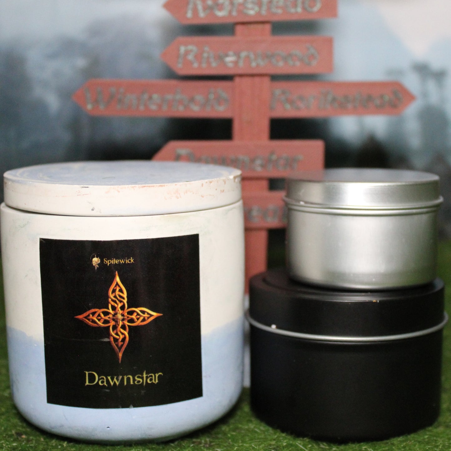 Front view of a handcrafted white and blue candle holder with a black label featuring a four pointed star in gold Celtic knotwork, with the name Dawnstar in gold below. Two smaller candles—one black 4oz and one silver 2oz—show the three available sizes. Set on grass in front of a 3D-printed signpost marking Skyrim cities, blending medieval fantasy with artisan craftsmanship.