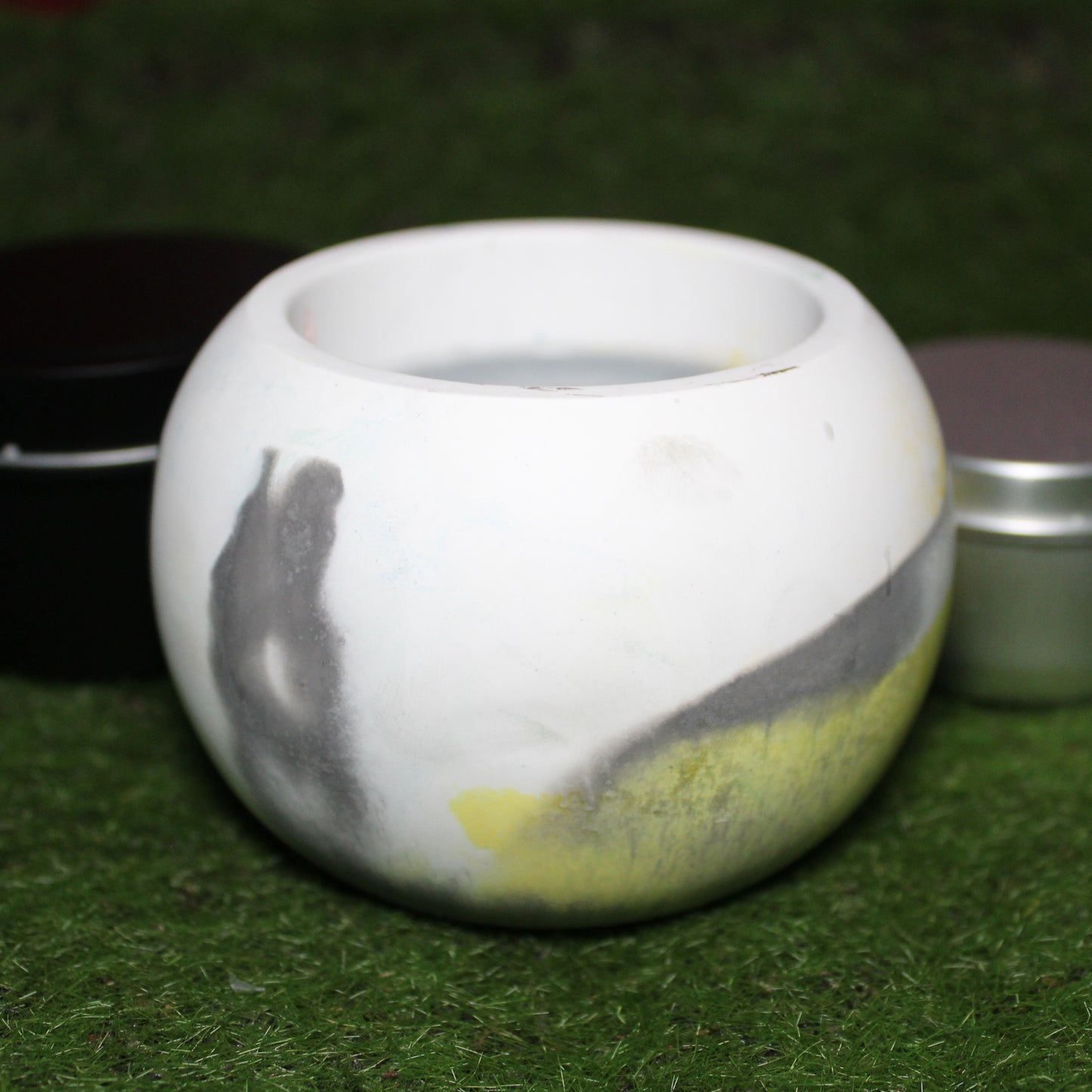Handcrafted Ecocrete candle holder featuring the grey white and yellow colors of the Demigender pride flag blended smoothly together. Product shot on grass with styling that includes a silver 2oz candle tin and black 4oz candle tin behind it, displaying the three available sizes of this LGBTQIA+ home decor piece.