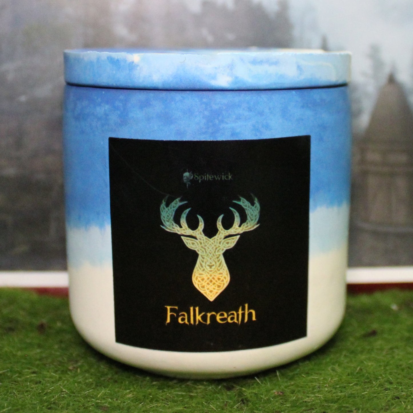 Front view of a handcrafted blue and white candle holder with a black label featuring a deer in gold Celtic knotwork. The name Falkreath is elegantly printed in gold beneath the design. Set on grass, this piece blends artisan craftsmanship with bold identity, making it perfect for unique home decor, gifts, and statement lighting.