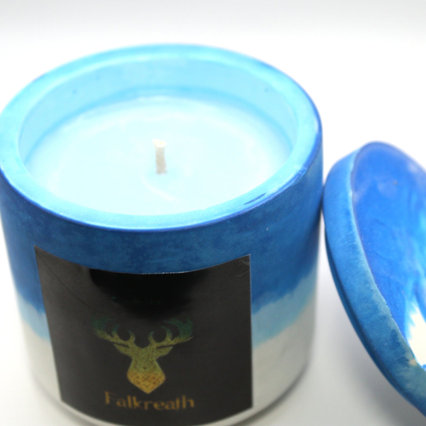 3/4 view of a blue and white handcrafted candle holder featuring a black label adorned with a deer in stylized gold Celtic knotwork. Beneath the intricate design, the name Falkreath is elegantly displayed in gold. The lid is removed and propped against the right side, revealing the blue candle inside. Set against a clean white background, this unique piece blends tradition with modern artistry, perfect for home decor, gifts, and personal expression.