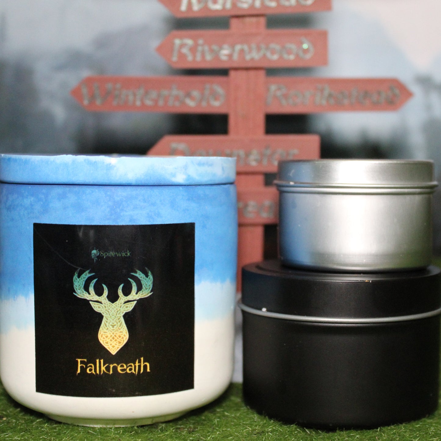 Front view of a handcrafted blue and white candle holder with a black label featuring a deer in gold Celtic knotwork, with the name Falkreath in gold below. Two smaller candlesâone black 4oz and one silver 2ozâshow the three available sizes. Set on grass in front of a 3D-printed signpost marking Skyrim cities, blending medieval fantasy with artisan craftsmanship.