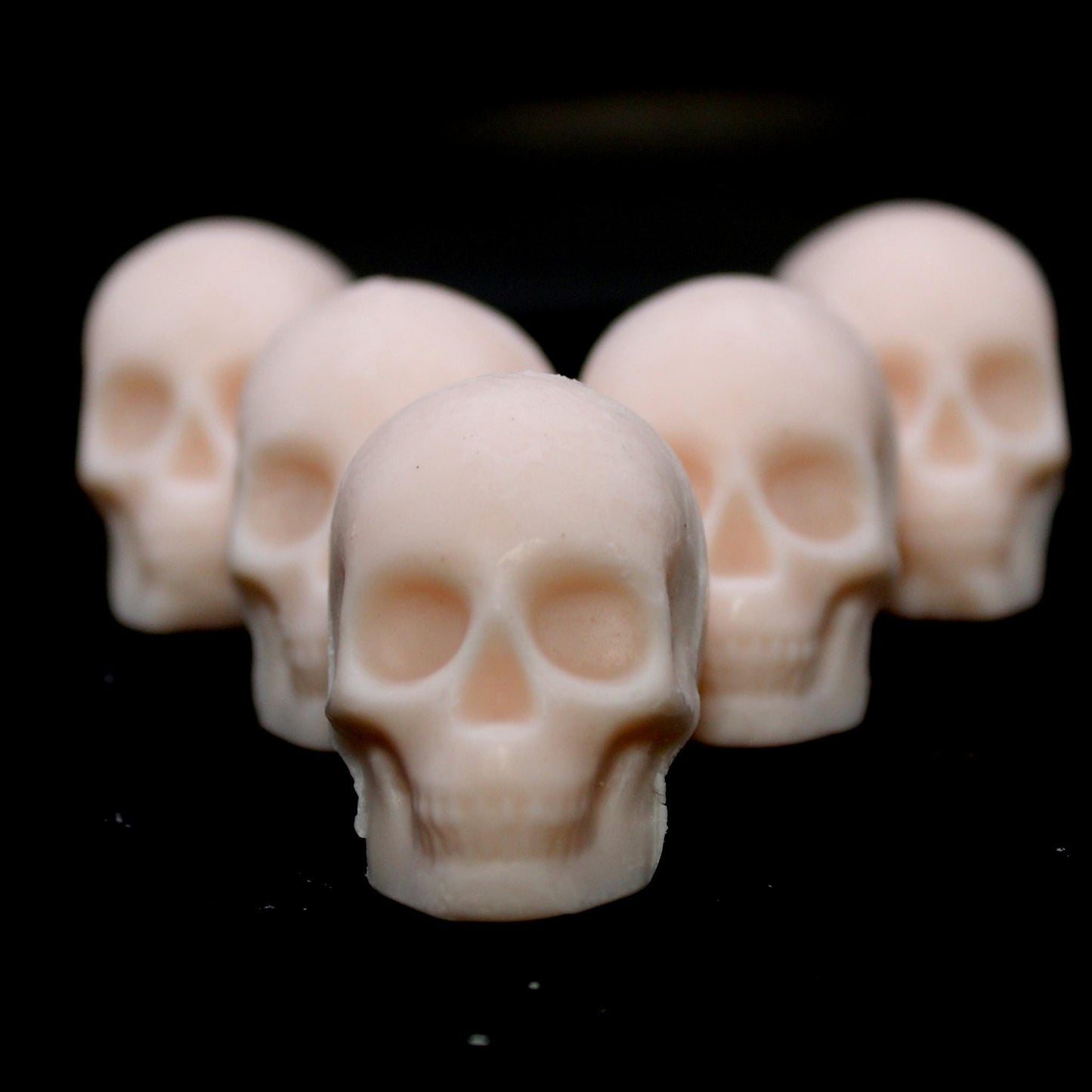 Five skull-shaped wax melts arranged in an arrowhead formation on a black background, each skull partially peeking from the one in front. The front skull stares directly into the camera, creating a bold, eerie aesthetic perfect for gothic decor, spooky ambiance, and macabre wax enthusiasts.