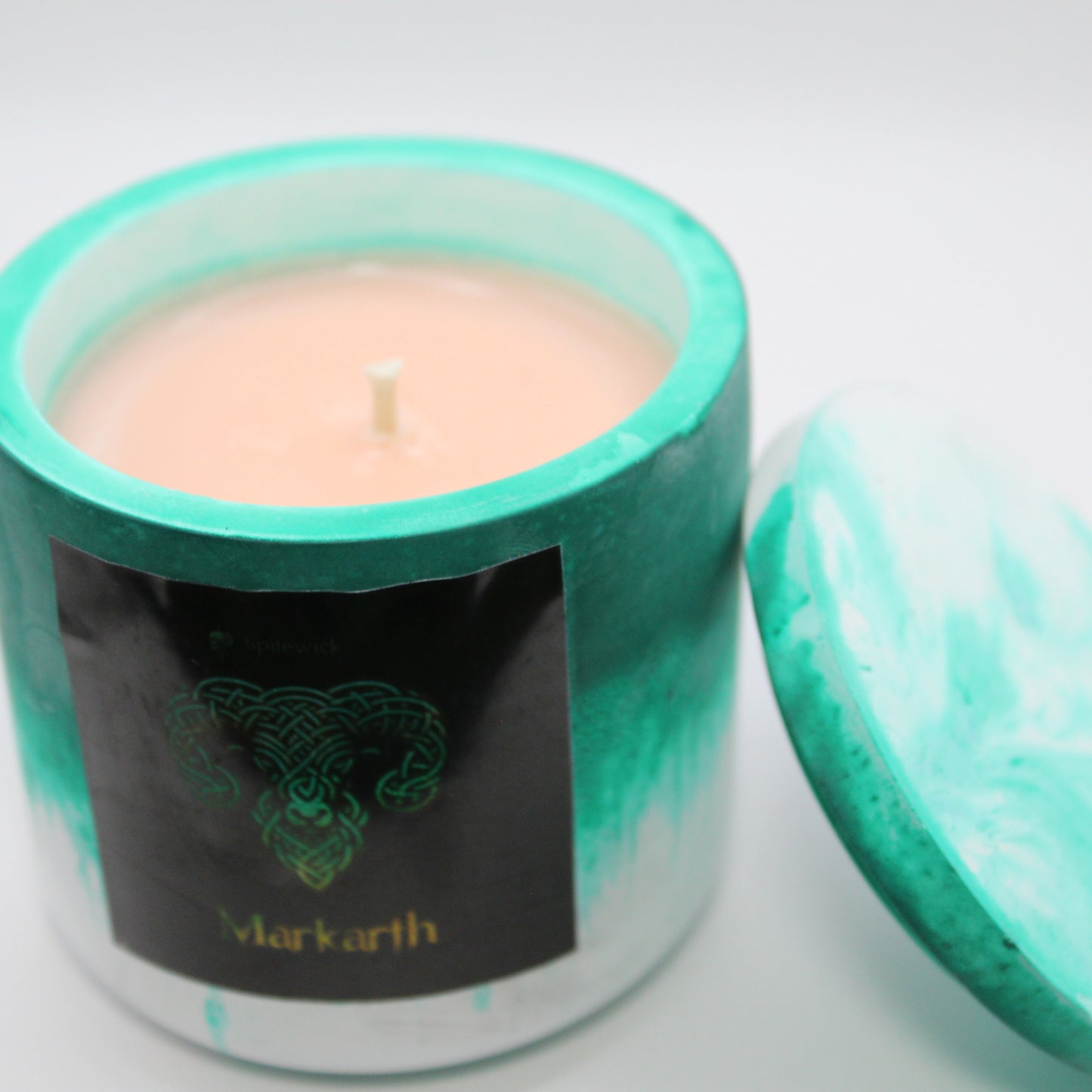 3/4 view of a green and white handcrafted candle holder featuring a black label adorned with a Ram in stylized gold Celtic knotwork. Beneath the intricate design, the name Markarth is elegantly displayed in gold. The lid is removed and propped against the right side, revealing the beige candle inside. Set against a clean white background, this unique piece blends tradition with modern artistry, perfect for home decor, gifts, and personal expression.