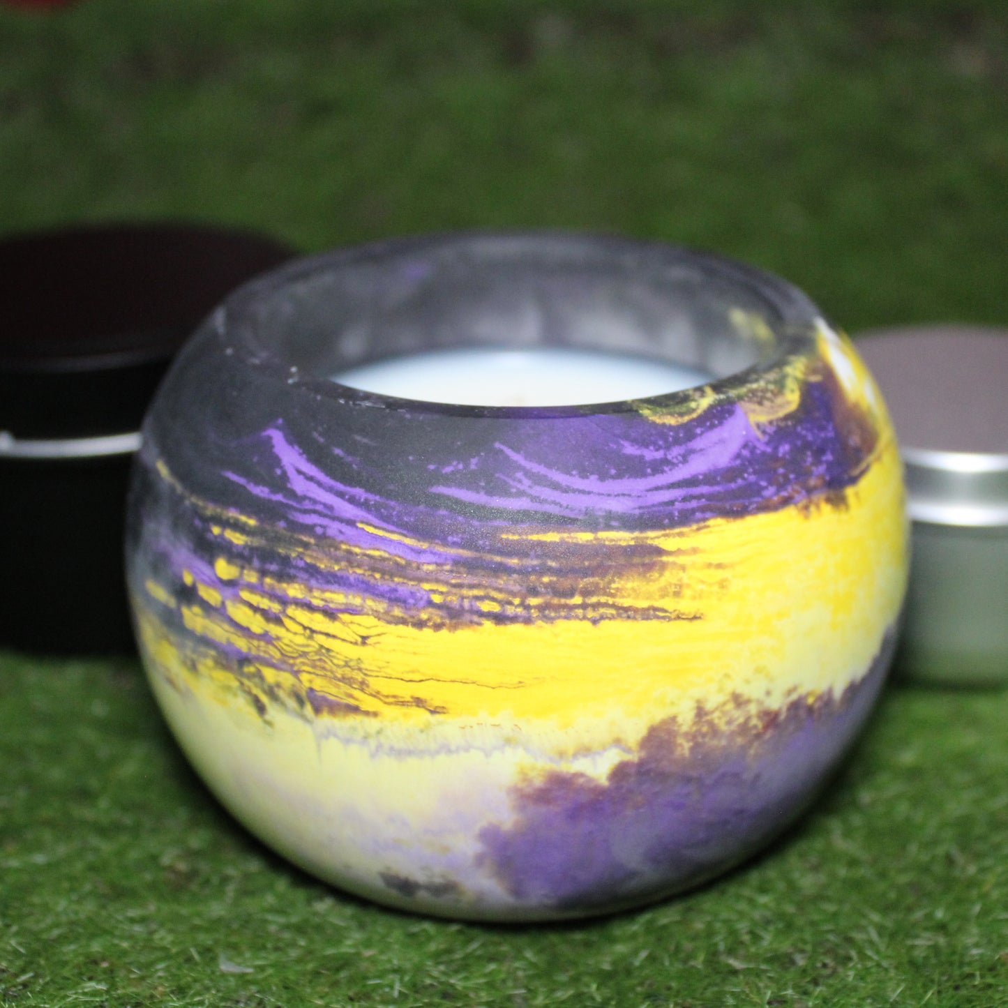Handcrafted Ecocrete candle holder featuring the black, purple, yellow and white colors of the nonbinary pride flag blended smoothly together. Product shot on grass with styling that includes a silver 2oz candle tin and black 4oz candle tin behind it, displaying the three available sizes of this LGBTQIA+ home decor piece.