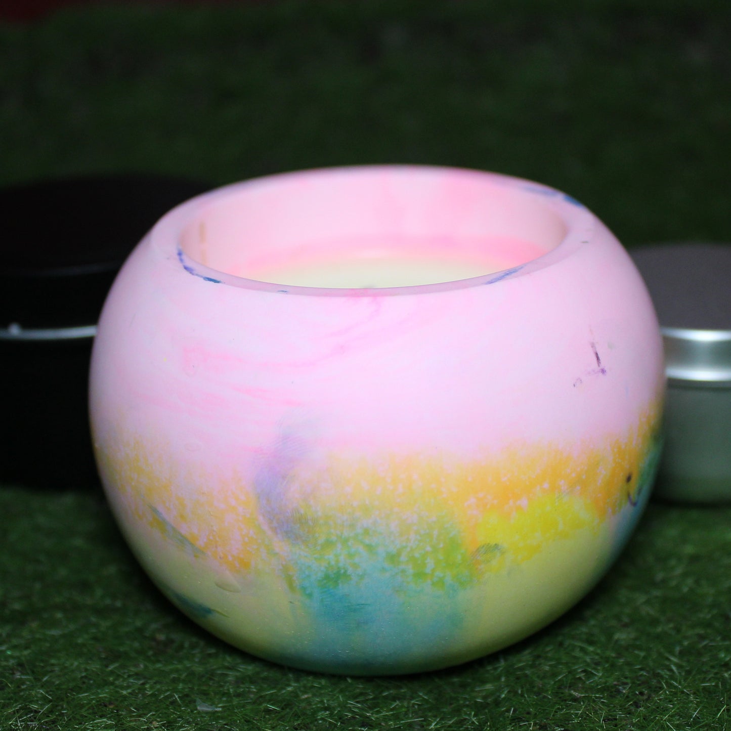 Handcrafted Ecocrete candle holder featuring the pink, yellow, and blue colors of the pansexual pride flag blended smoothly together. Product shot on natural grass with styling that includes a silver 2oz candle tin and black 4oz candle tin behind it, displaying the three available sizes of this LGBTQIA+ home decor piece.