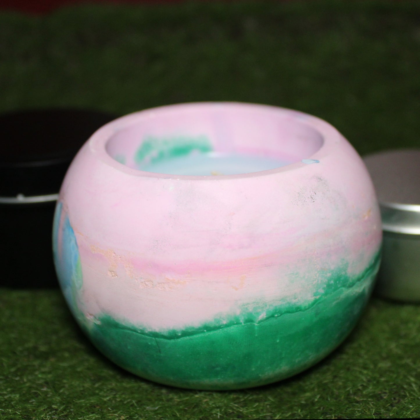 Handcrafted Ecocrete candle holder featuring the blue pink and green colors of the polysexual pride flag blended smoothly together. Product shot on natural grass with styling that includes a silver 2oz candle tin and black 4oz candle tin behind it, displaying the three available sizes of this LGBTQIA+ home decor piece.