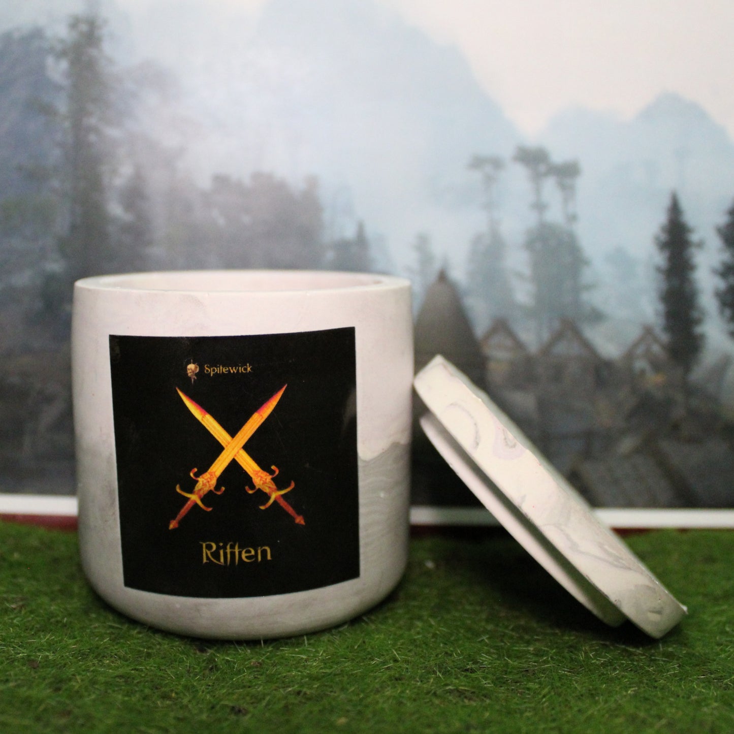 Front view of a handcrafted soft purple and grey candle holder with a black label featuring two crossed swords in gold Celtic knotwork. The name riften is elegantly printed in gold beneath the design. Set on grass, this piece blends artisan craftsmanship with bold identity, making it perfect for unique home decor, gifts, and statement lighting.