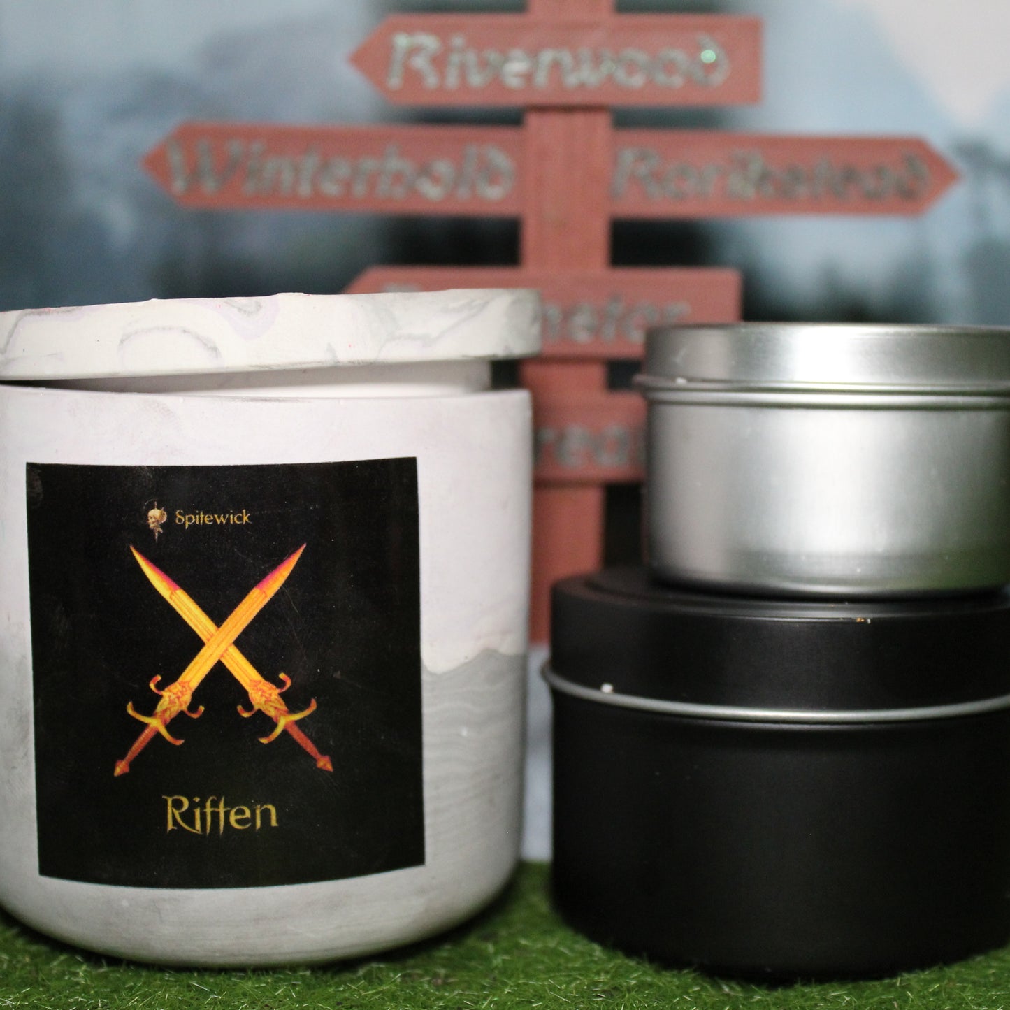 Front view of a handcrafted soft purple and grey candle holder with a black label featuring two crossed swors in gold Celtic knotwork, with the name Riften in gold below. Two smaller candles—one black 4oz and one silver 2oz—show the three available sizes. Set on grass in front of a 3D-printed signpost marking Skyrim cities, blending medieval fantasy with artisan craftsmanship.
