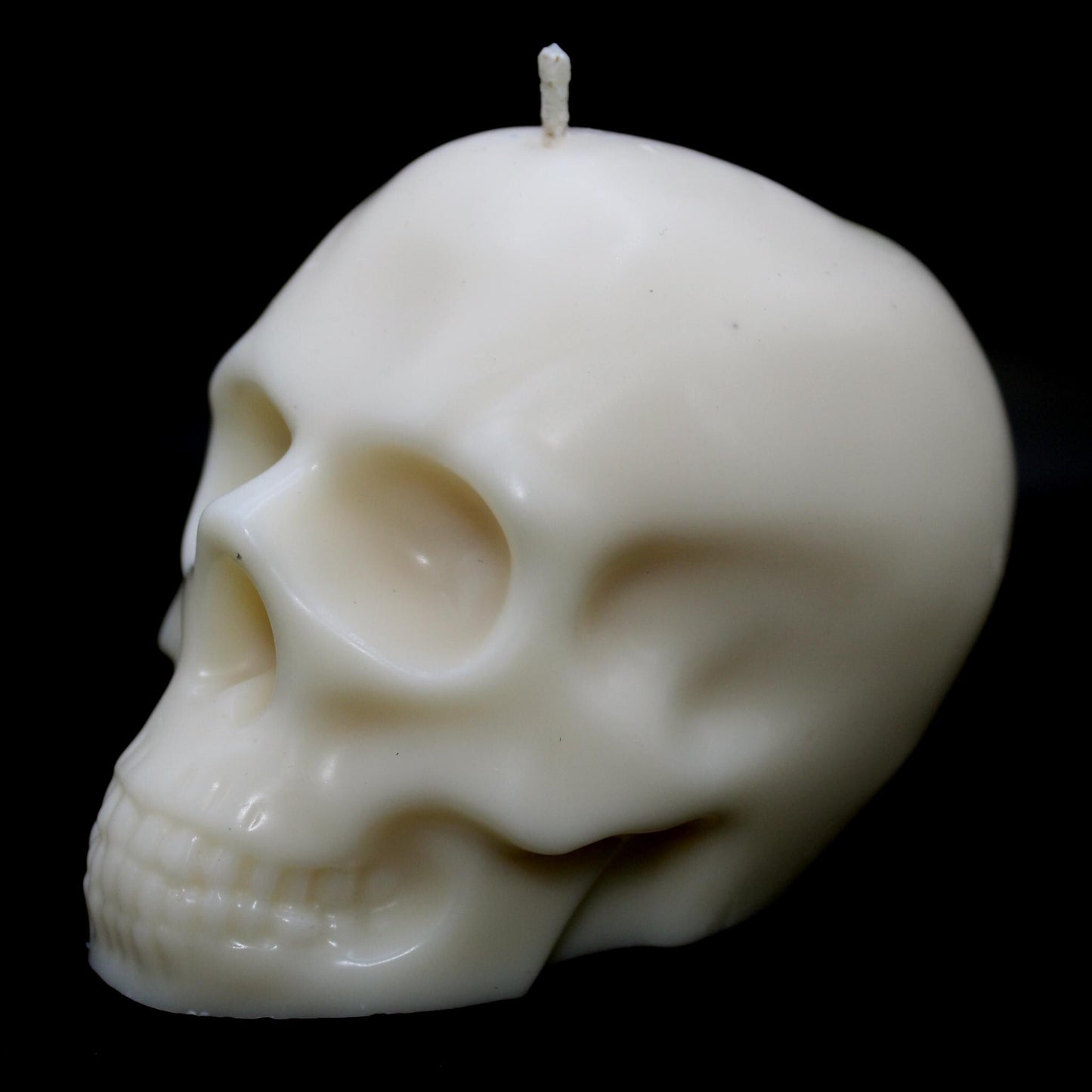 White skull-shaped wax candle against a black background, turned slightly to the left, highlighting intricate details on the right side. A bold gothic decor piece perfect for dark aesthetic lovers, spooky home accents, and macabre candle enthusiasts.