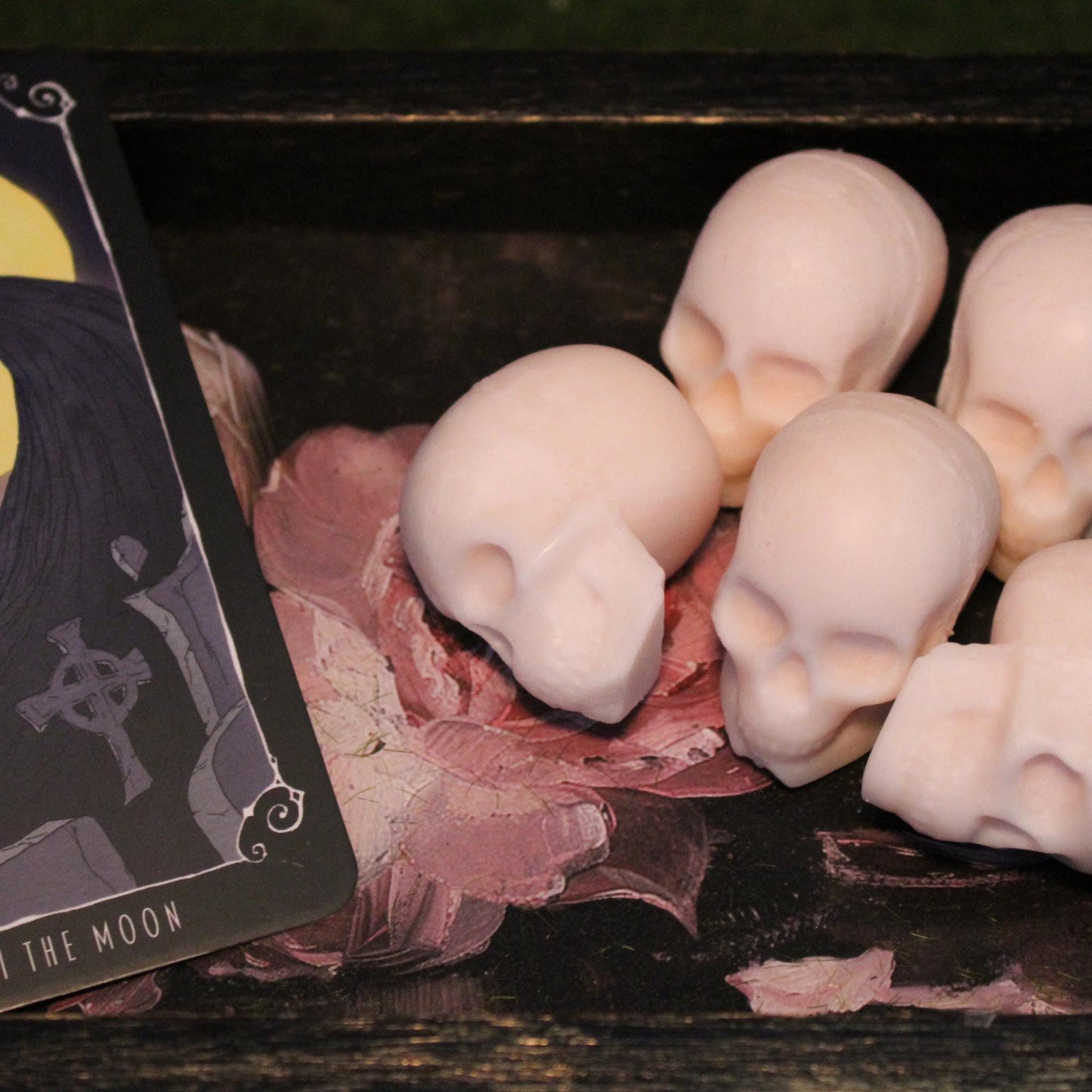 Five skull-shaped wax melts resting on an antique tray surface with a cracked painted flower design and faded gold gilding. The corner of 'The Moon' tarot card is visible, adding a mystical, gothic aesthetic. Perfect for dark decor, occult-themed spaces, and macabre candle lovers.