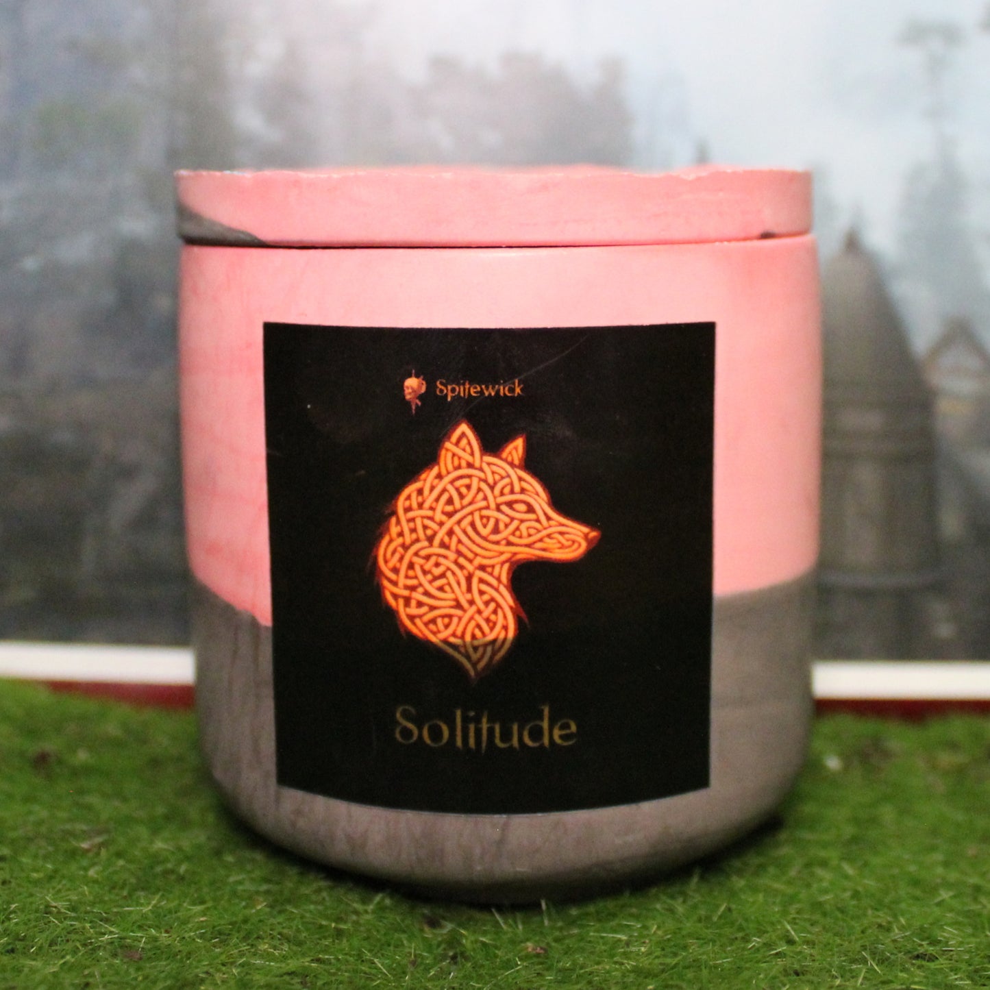 Front view of a handcrafted red and black candle holder with a black label featuring a wolf in gold Celtic knotwork. The name Solitude is elegantly printed in gold beneath the design. Set on grass, this piece blends artisan craftsmanship with bold identity, making it perfect for unique home decor, gifts, and statement lighting.
