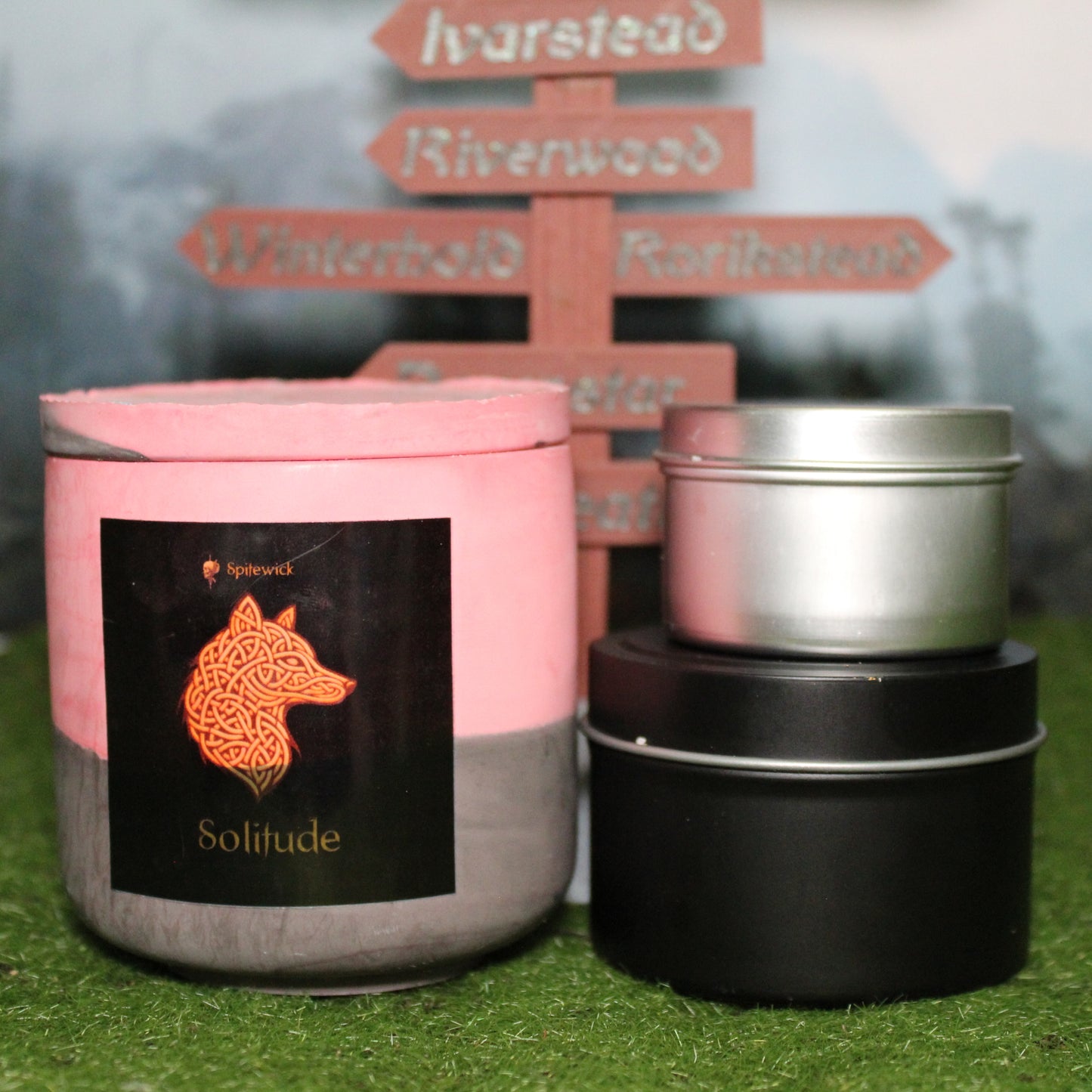 Front view of a handcrafted red and black candle holder with a black label featuring a wofl in gold Celtic knotwork, with the name solitude in gold below. Two smaller candlesâone black 4oz and one silver 2ozâshow the three available sizes. Set on grass in front of a 3D-printed signpost marking Skyrim cities, blending medieval fantasy with artisan craftsmanship.