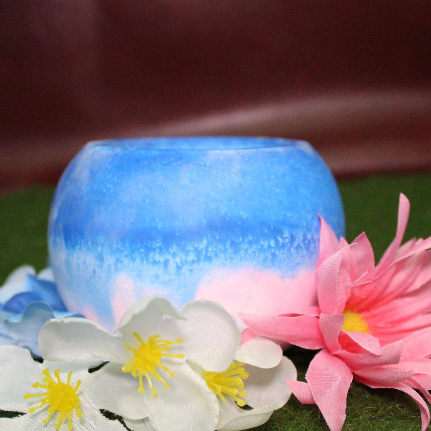 Handcrafted candle holder featuring the pink, blue, and white hues of the transgender pride flag, seamlessly blended together for a striking design. Set on grass with a burgundy vinyl backdrop, surrounded by an assortment of flowers mirroring the flag’s colors, creating a visually rich and identity-affirming scene. Perfect for LGBTQ+ home decor, pride gifts, and statement lighting."