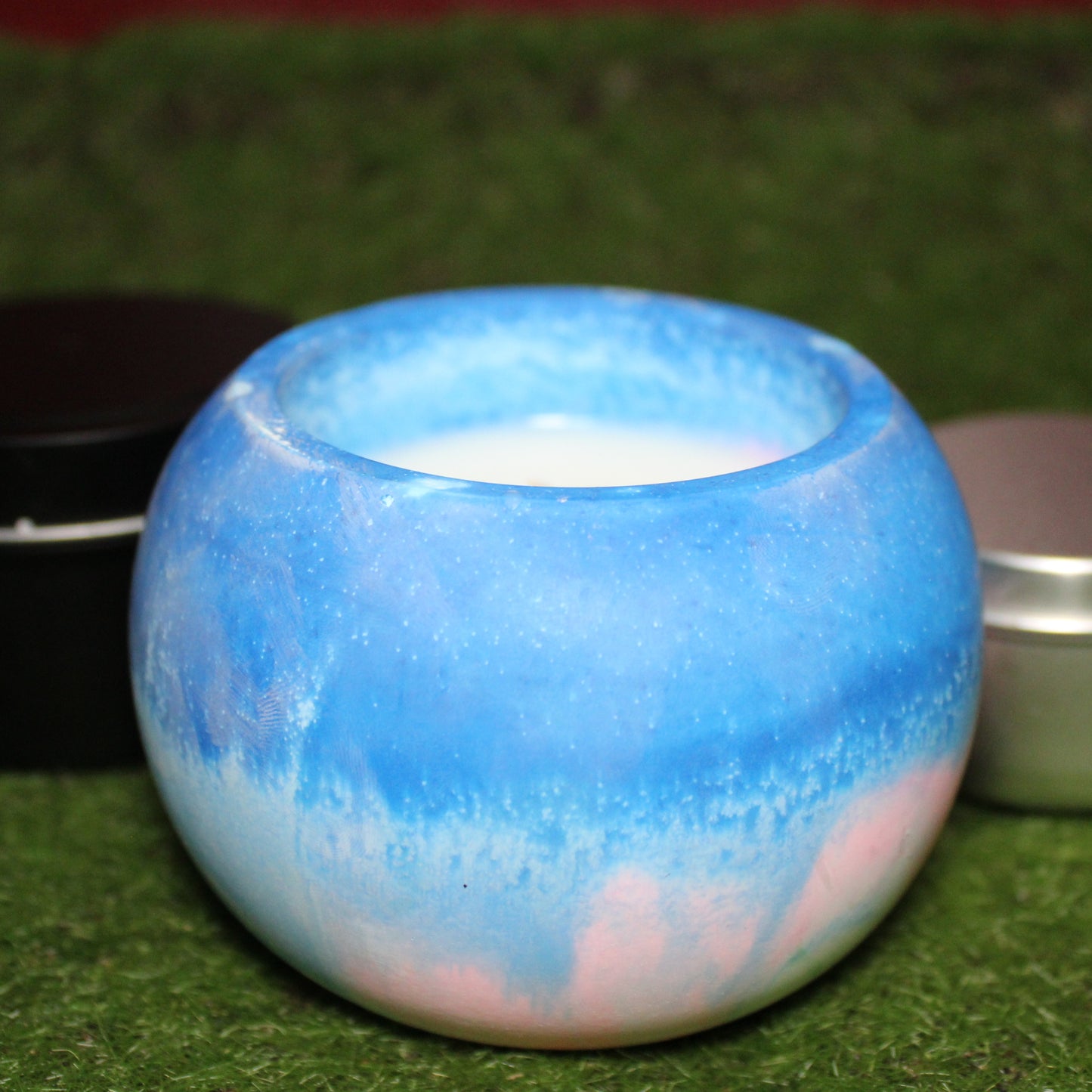 Handcrafted ecocrete candle holder featuring the blue, pink, and white colors of the transgender pride flag blended smoothly together. Product shot on grass with styling that includes a silver 2oz candle tin and black 4oz candle tin behind it, displaying the three available sizes of this LGBTQIA+ home decor piece.
