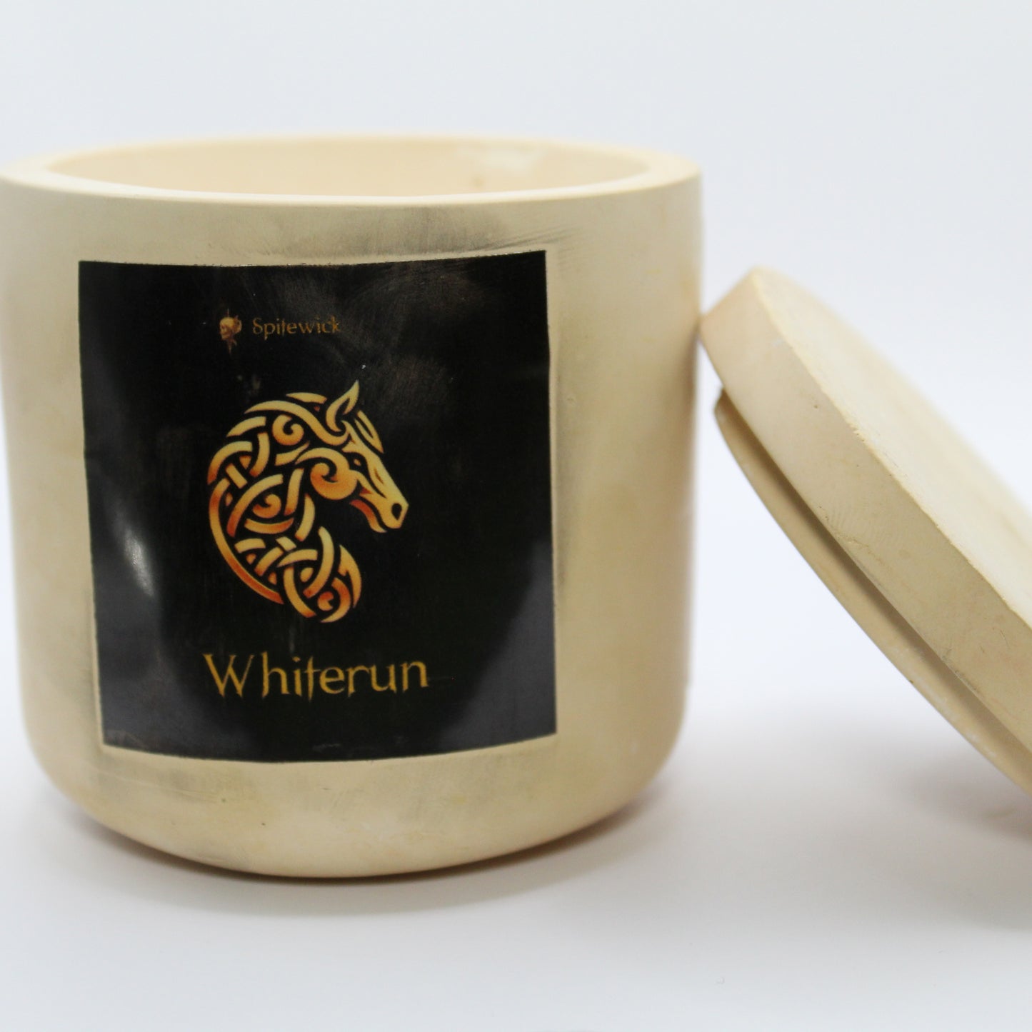 A beige candle holder with a black label featuring a horse in elegant gold Celtic knotwork. The name whiterun is displayed in gold beneath the intricate design, adding a bold, mystical touch. The lid is removed and propped against the right side, showcasing the full aesthetic. Set against a clean white background, this handcrafted candle merges tradition and modern artistry, perfect for unique home decor, gifts, and personal expression.