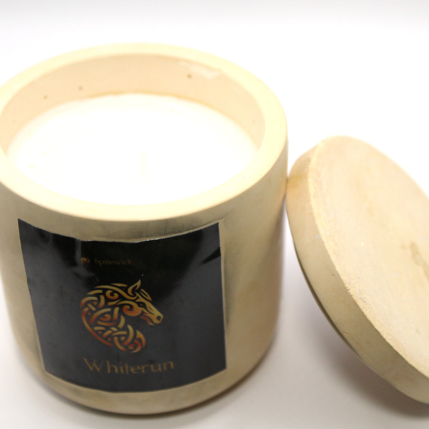 3/4 view of a beige handcrafted candle holder featuring a black label adorned with a horse in stylized gold Celtic knotwork. Beneath the intricate design, the name whiterun is elegantly displayed in gold. The lid is removed and propped against the right side, revealing the <color> candle inside. Set against a clean white background, this unique piece blends tradition with modern artistry, perfect for home decor, gifts, and personal expression.