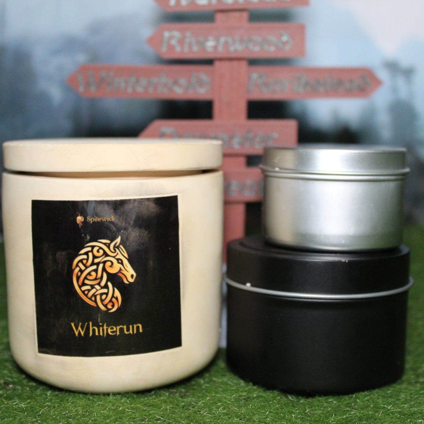 Front view of a handcrafted beige candle holder with a black label featuring a horse in gold Celtic knotwork, with the name whiterun in gold below. Two smaller candlesâone black 4oz and one silver 2ozâshow the three available sizes. Set on grass in front of a 3D-printed signpost marking Skyrim cities, blending medieval fantasy with artisan craftsmanship.