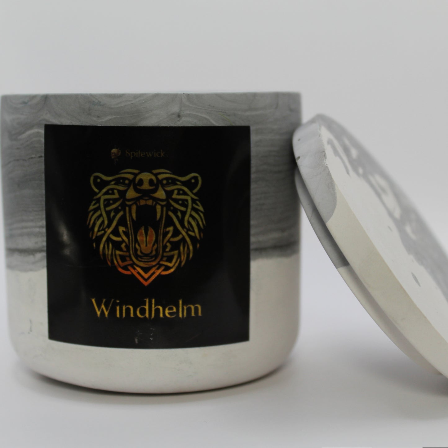 A black and white candle holder with a black label featuring a bear in elegant gold Celtic knotwork. The name windhelm is displayed in gold beneath the intricate design, adding a bold, mystical touch. The lid is removed and propped against the right side, showcasing the full aesthetic. Set against a clean white background, this handcrafted candle merges tradition and modern artistry, perfect for unique home decor, gifts, and personal expression.