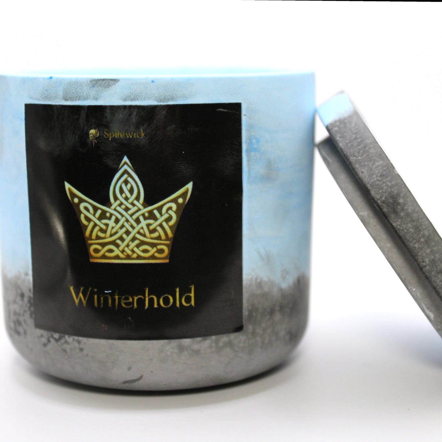 A blue and black candle holder with a black label featuring a Crown in elegant gold Celtic knotwork. The name Winterhold is displayed in gold beneath the intricate design, adding a bold, mystical touch. The lid is removed and propped against the right side, showcasing the full aesthetic. Set against a clean white background, this handcrafted candle merges tradition and modern artistry, perfect for unique home decor, gifts, and personal expression.