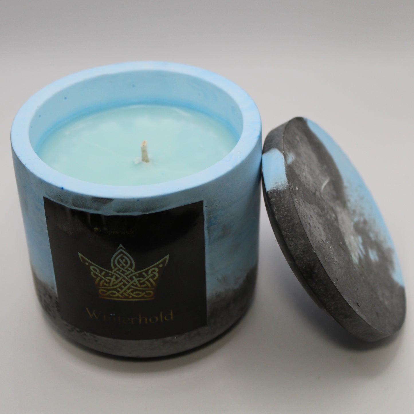 3/4 view of a blue and black handcrafted candle holder featuring a black label adorned with a Crown in stylized gold Celtic knotwork. Beneath the intricate design, the name Winterhold is elegantly displayed in gold. The lid is removed and propped against the right side, revealing the blue candle inside. Set against a clean white background, this unique piece blends tradition with modern artistry, perfect for home decor, gifts, and personal expression.