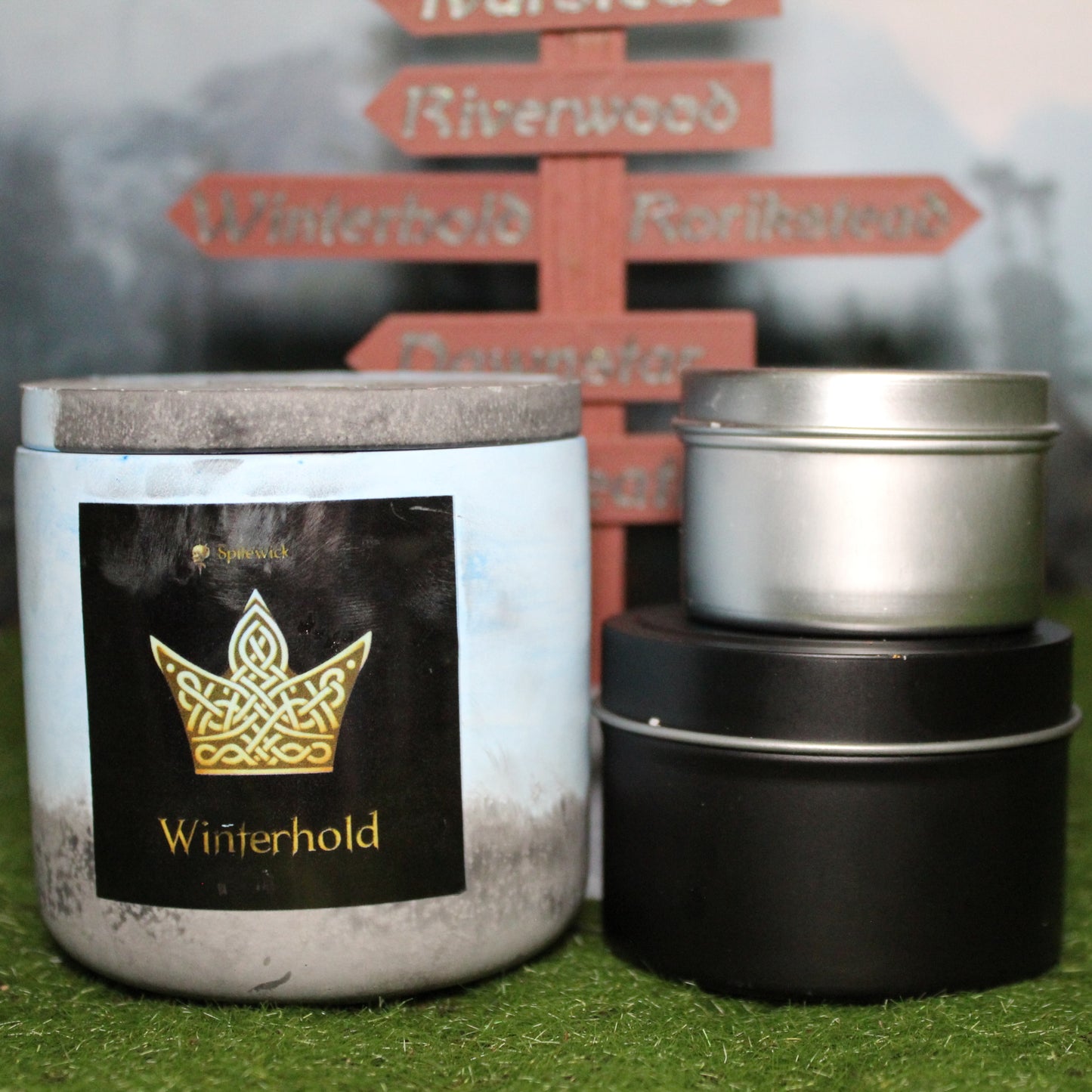 Front view of a handcrafted blue and black candle holder with a black label featuring a Crown in gold Celtic knotwork, with the name winterhold in gold below. Two smaller candles—one black 4oz and one silver 2oz—show the three available sizes. Set on grass in front of a 3D-printed signpost marking Skyrim cities, blending medieval fantasy with artisan craftsmanship.