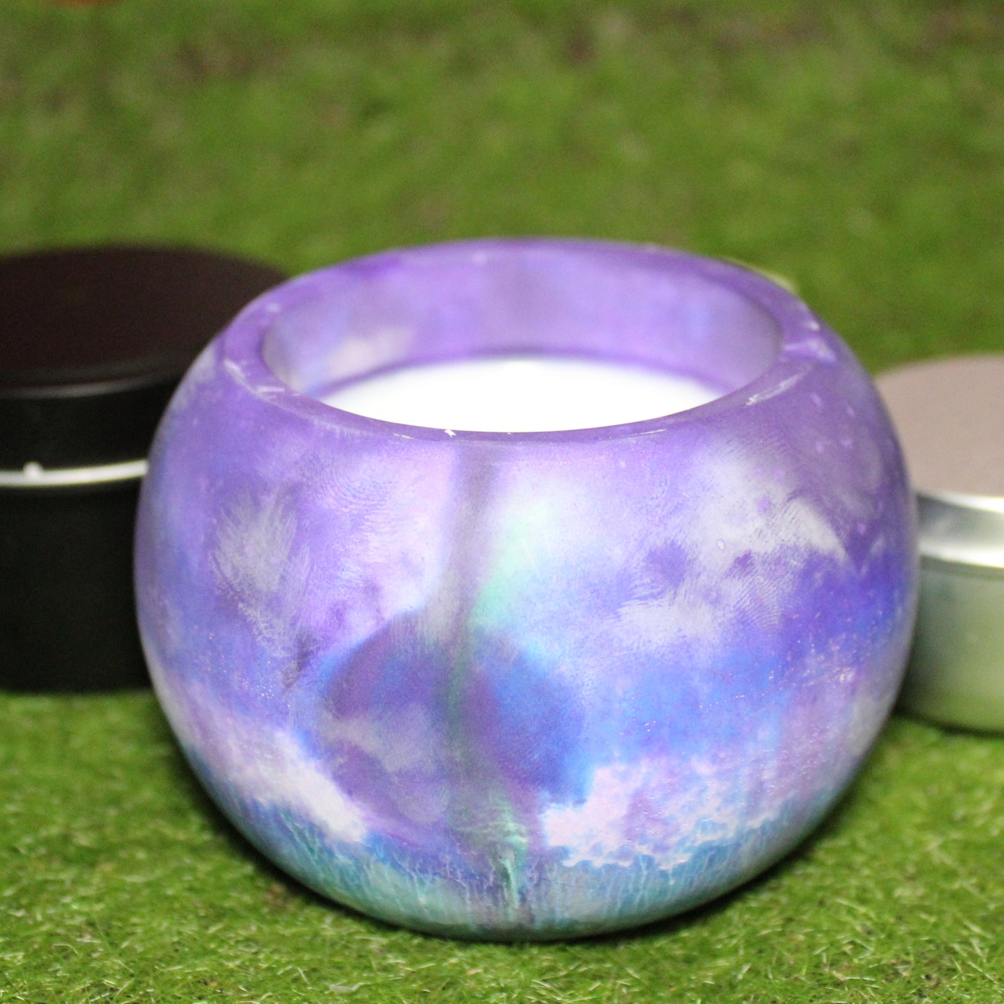 Handcrafted Ecocrete candle holder featuring the purple, green, blue and white colors of the Trans-Inclusive Gay Men's pride flag blended smoothly together. Product shot on grass with styling that includes a silver 2oz candle tin and black 4oz candle tin behind it, displaying the three available sizes of this LGBTQIA+ home decor piece.