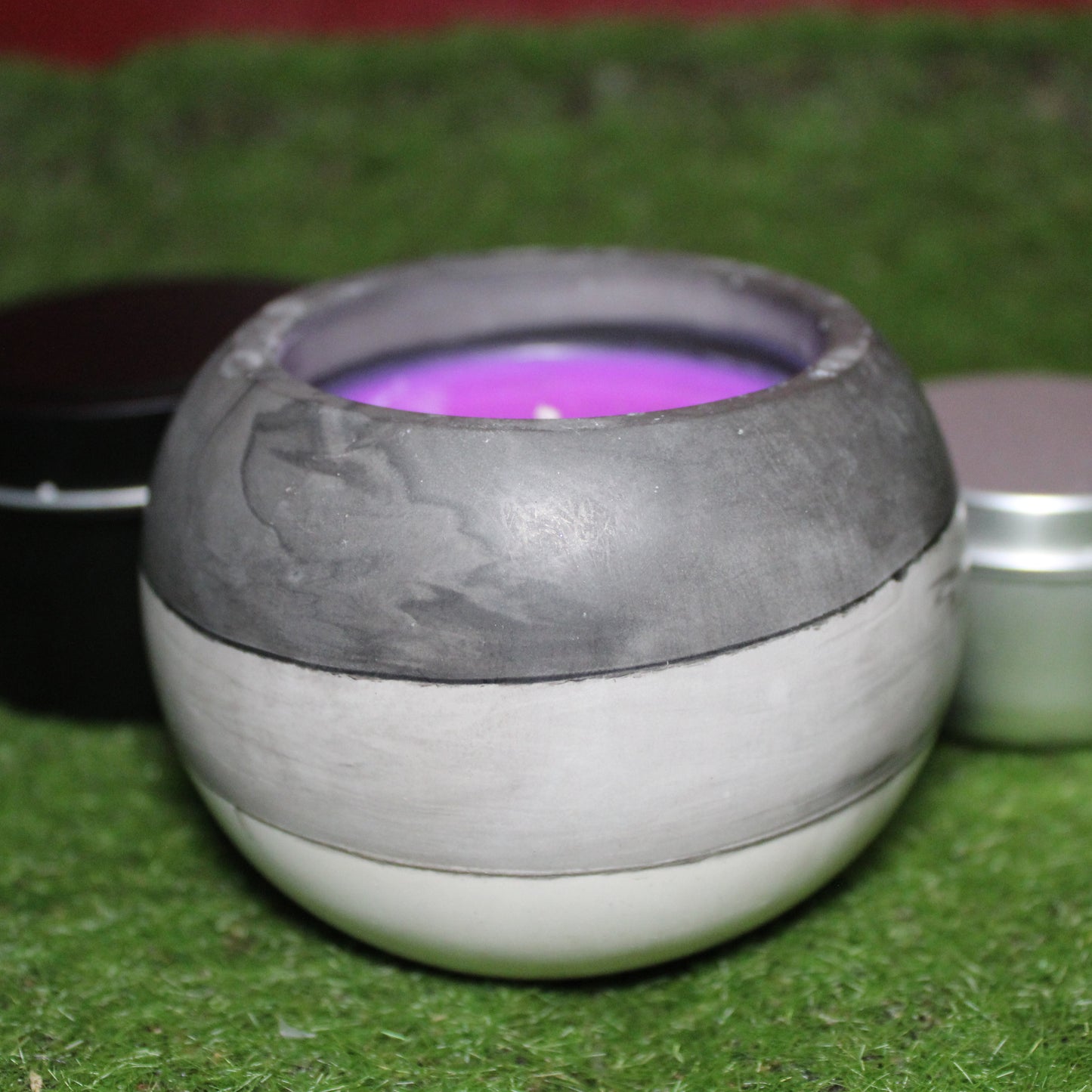 Handcrafted Ecocrete candle holder featuring the black, white and grey colors of the asexual pride flag blended smoothly together. Product shot on grass with styling that includes a silver 2oz candle tin and black 4oz candle tin behind it, displaying the three available sizes of this LGBTQIA+ home decor piece.