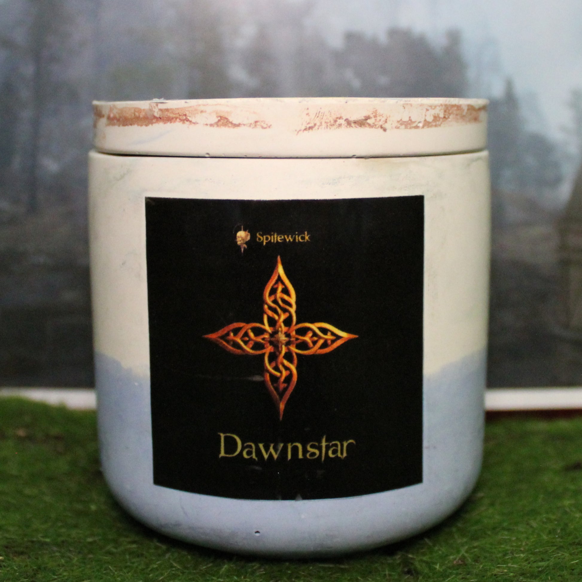 Front view of a handcrafted white and pale blue candle holder with a black label featuring a four pointed star in gold Celtic knotwork. The name dawnstar is elegantly printed in gold beneath the design. Set on grass, this piece blends artisan craftsmanship with bold identity, making it perfect for unique home decor, gifts, and statement lighting.