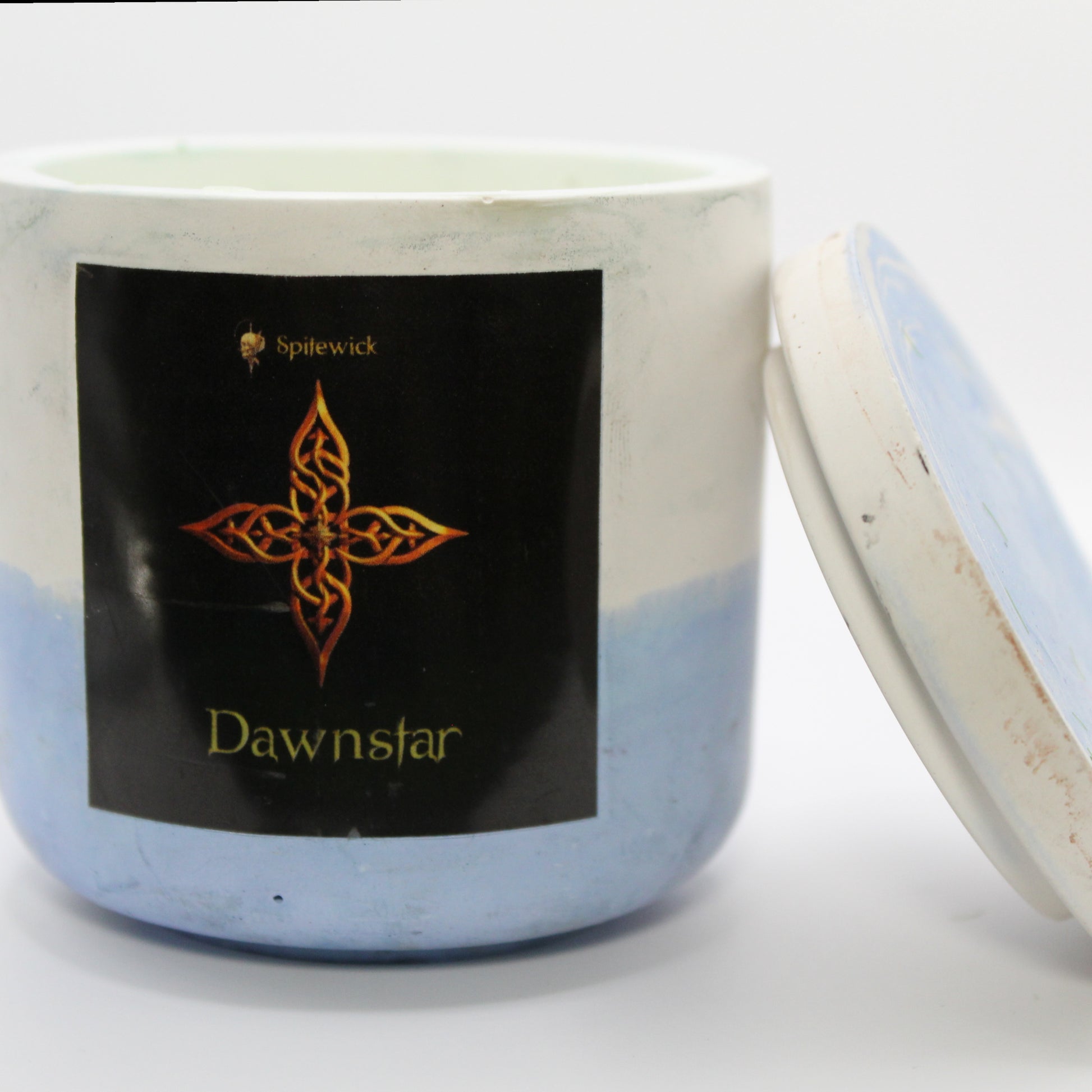 A white and blue candle holder with a black label featuring a four-pointed star in elegant gold Celtic knotwork. The name dawnstar is displayed in gold beneath the intricate design, adding a bold, mystical touch. The lid is removed and propped against the right side, showcasing the full aesthetic. Set against a clean white background, this handcrafted candle merges tradition and modern artistry, perfect for unique home decor, gifts, and personal expression.