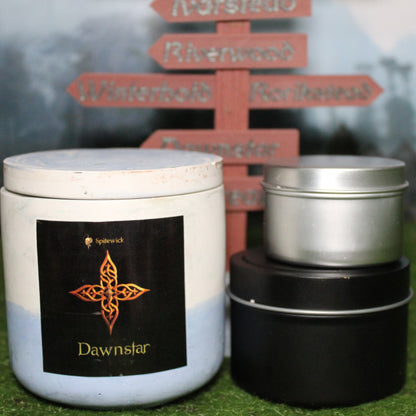 Front view of a handcrafted white and blue candle holder with a black label featuring a four pointed star in gold Celtic knotwork, with the name Dawnstar in gold below. Two smaller candles—one black 4oz and one silver 2oz—show the three available sizes. Set on grass in front of a 3D-printed signpost marking Skyrim cities, blending medieval fantasy with artisan craftsmanship.