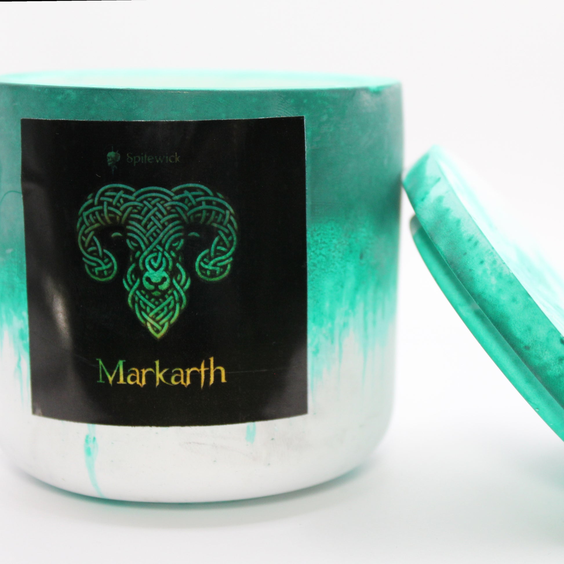 A green and white candle holder with a black label featuring a ram in elegant gold Celtic knotwork. The name Markarth is displayed in gold beneath the intricate design, adding a bold, mystical touch. The lid is removed and propped against the right side, showcasing the full aesthetic. Set against a clean white background, this handcrafted candle merges tradition and modern artistry, perfect for unique home decor, gifts, and personal expression.
