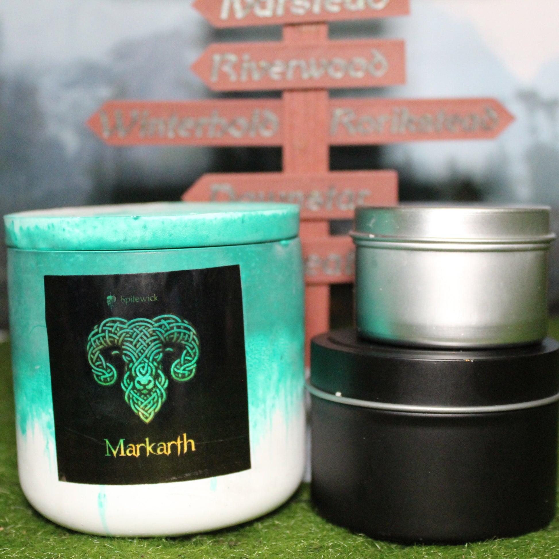 Front view of a handcrafted green and white candle holder with a black label featuring a ram in gold Celtic knotwork, with the name Markarth in gold below. Two smaller candles—one black 4oz and one silver 2oz—show the three available sizes. Set on grass in front of a 3D-printed signpost marking Skyrim cities, blending medieval fantasy with artisan craftsmanship.