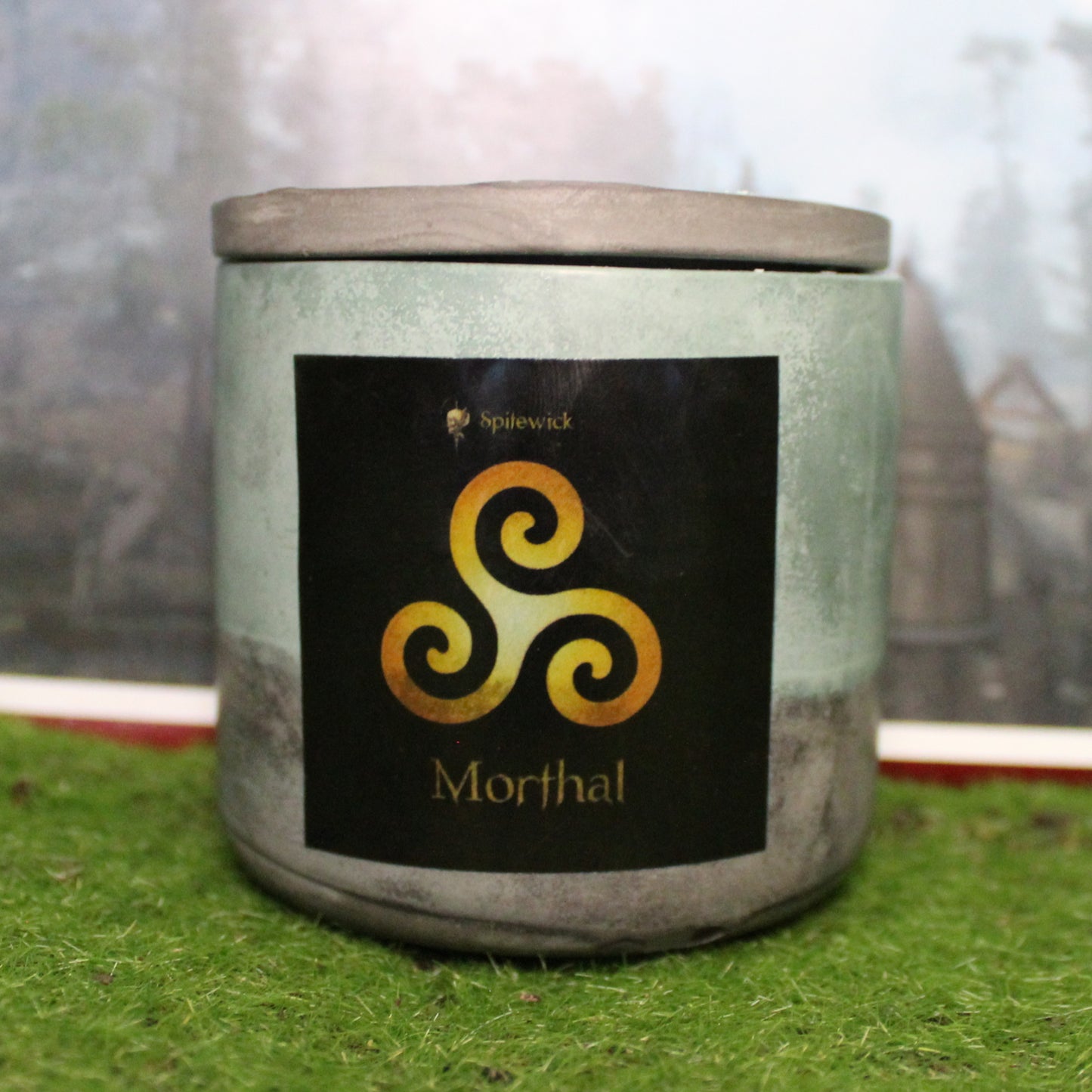 Front view of a handcrafted green and black candle holder with a black label featuring a Triskele in gold. The name Morthal is elegantly printed in gold beneath the design. Set on grass, this piece blends artisan craftsmanship with bold identity, making it perfect for unique home decor, gifts, and statement lighting.