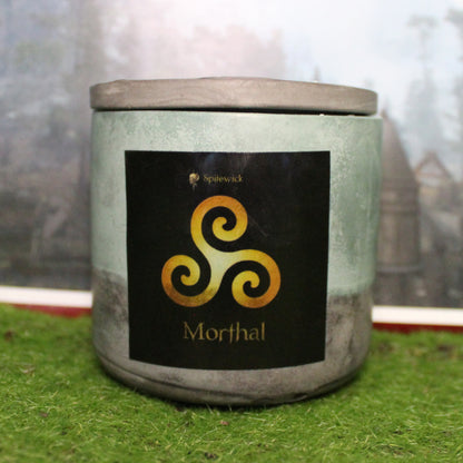 Front view of a handcrafted green and black candle holder with a black label featuring a Triskele in gold. The name Morthal is elegantly printed in gold beneath the design. Set on grass, this piece blends artisan craftsmanship with bold identity, making it perfect for unique home decor, gifts, and statement lighting.