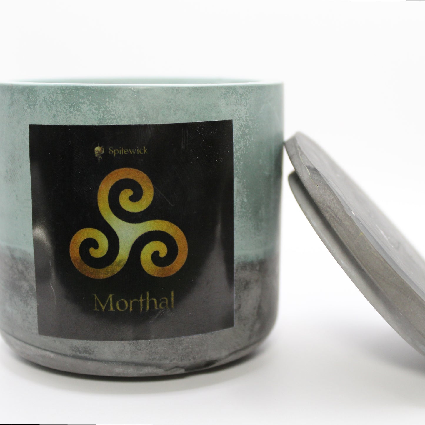 A green and black candle holder with a black label featuring a Triskele in elegant gold. The name Morthal is displayed in gold beneath the intricate design, adding a bold, mystical touch. The lid is removed and propped against the right side, showcasing the full aesthetic. Set against a clean white background, this handcrafted candle merges tradition and modern artistry, perfect for unique home decor, gifts, and personal expression.