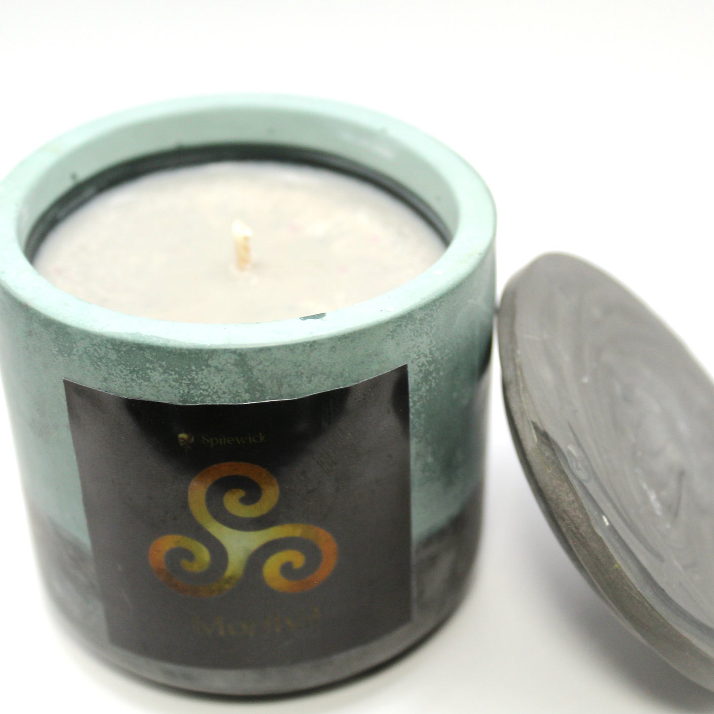 3/4 view of a green and black handcrafted candle holder featuring a black label adorned with a Triskele in stylized gold. Beneath the intricate design, the name Morthal is elegantly displayed in gold. The lid is removed and propped against the right side, revealing the white candle inside. Set against a clean white background, this unique piece blends tradition with modern artistry, perfect for home decor, gifts, and personal expression.