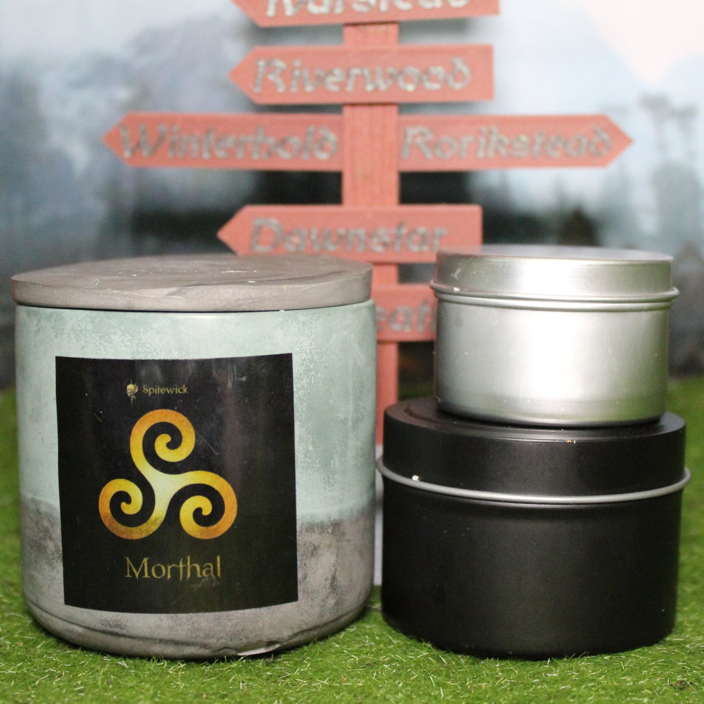 Front view of a handcrafted green and black candle holder with a black label featuring a Triskele in gold, with the name Morthal in gold below. Two smaller candles—one black 4oz and one silver 2oz—show the three available sizes. Set on grass in front of a 3D-printed signpost marking Skyrim cities, blending medieval fantasy with artisan craftsmanship.