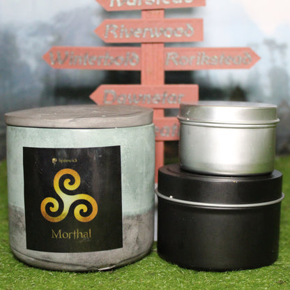Front view of a handcrafted green and black candle holder with a black label featuring a Triskele in gold, with the name Morthal in gold below. Two smaller candles—one black 4oz and one silver 2oz—show the three available sizes. Set on grass in front of a 3D-printed signpost marking Skyrim cities, blending medieval fantasy with artisan craftsmanship.