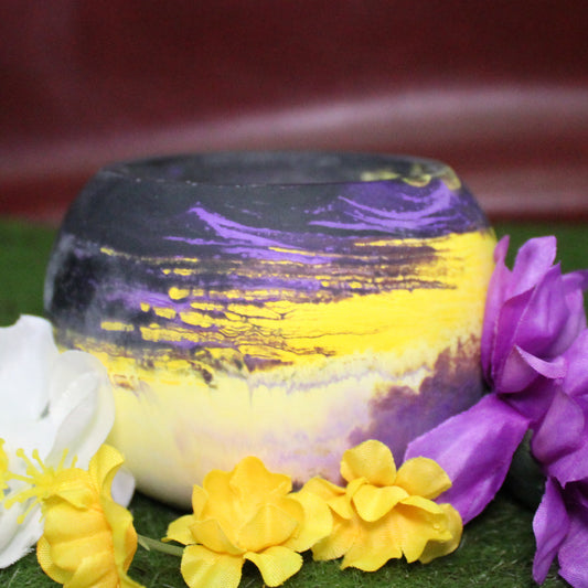 Handcrafted candle holder featuring the black, purple, yellow and white hues of the nonbinary pride flag, seamlessly blended together for a striking design. Set on grass with a burgundy vinyl backdrop, surrounded by an assortment of flowers mirroring the flag’s colors, creating a visually rich and identity-affirming scene. Perfect for LGBTQ+ home decor, pride gifts, and statement lighting.