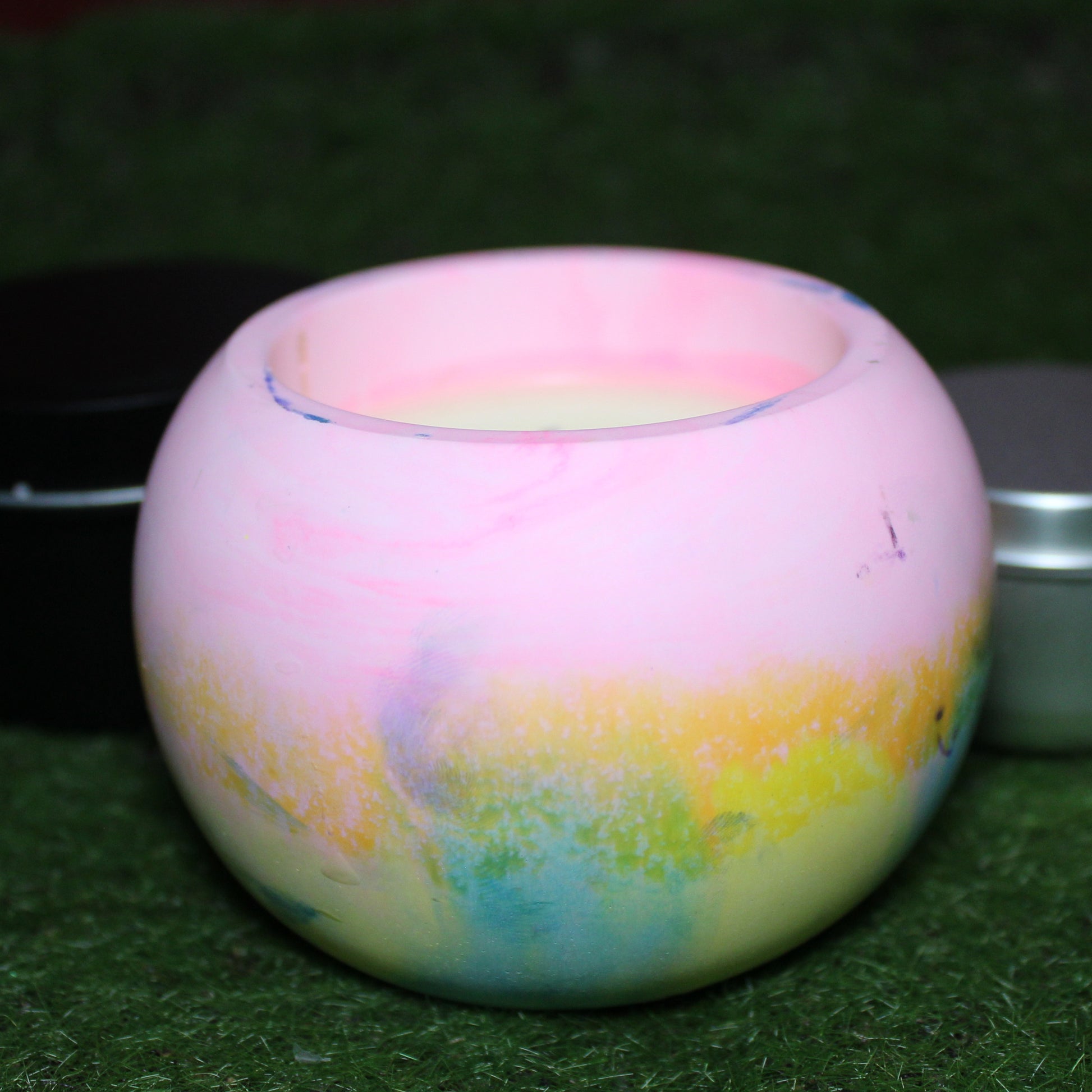 Handcrafted Ecocrete candle holder featuring the pink, yellow, and blue colors of the pansexual pride flag blended smoothly together. Product shot on natural grass with styling that includes a silver 2oz candle tin and black 4oz candle tin behind it, displaying the three available sizes of this LGBTQIA+ home decor piece.