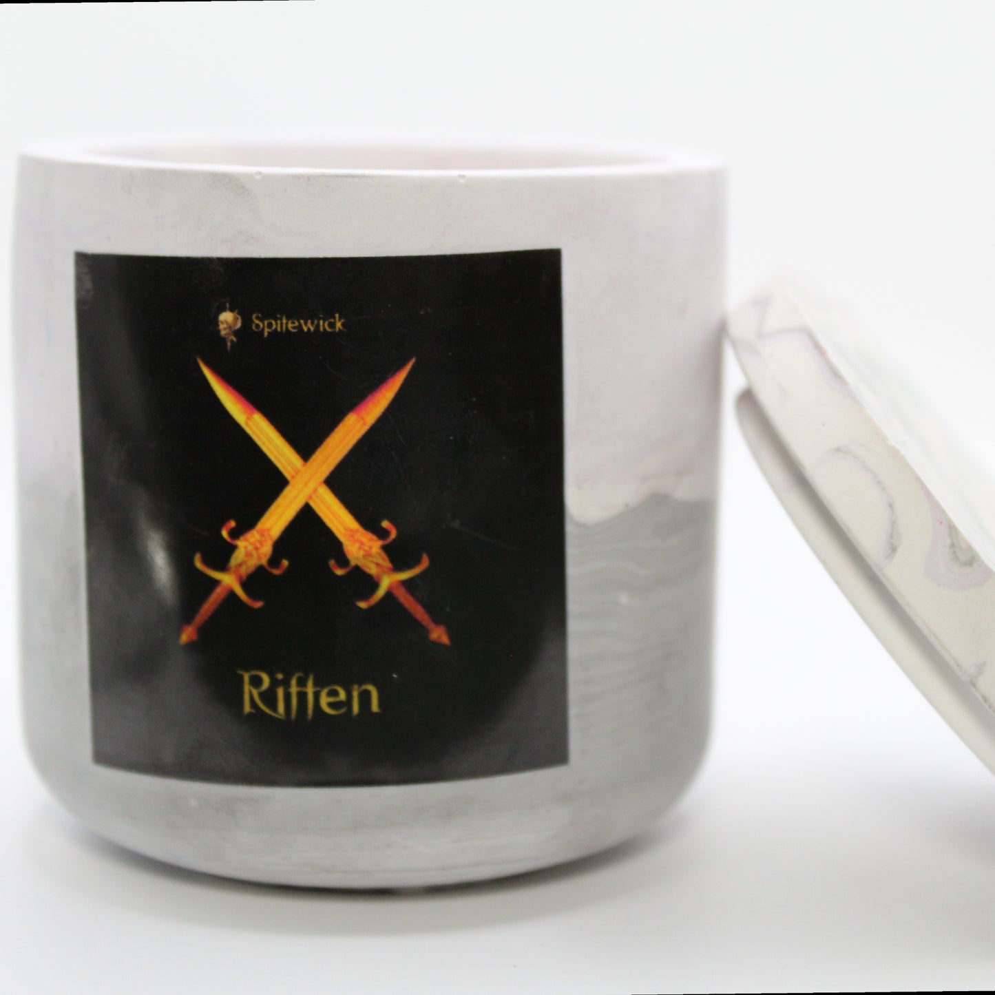 A soft purple and grey candle holder with a black label featuring a two crossed swords in elegant gold Celtic knotwork. The name Riften is displayed in gold beneath the intricate design, adding a bold, mystical touch. The lid is removed and propped against the right side, showcasing the full aesthetic. Set against a clean white background, this handcrafted candle merges tradition and modern artistry, perfect for unique home decor, gifts, and personal expression.