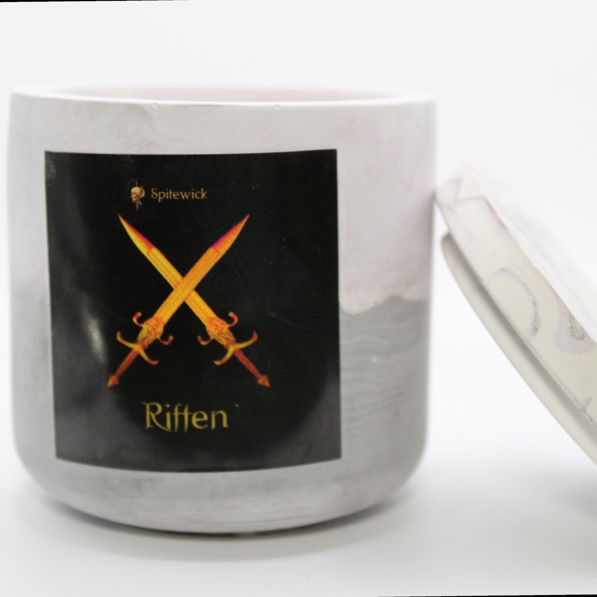A soft purple and grey candle holder with a black label featuring a two crossed swords in elegant gold Celtic knotwork. The name Riften is displayed in gold beneath the intricate design, adding a bold, mystical touch. The lid is removed and propped against the right side, showcasing the full aesthetic. Set against a clean white background, this handcrafted candle merges tradition and modern artistry, perfect for unique home decor, gifts, and personal expression.