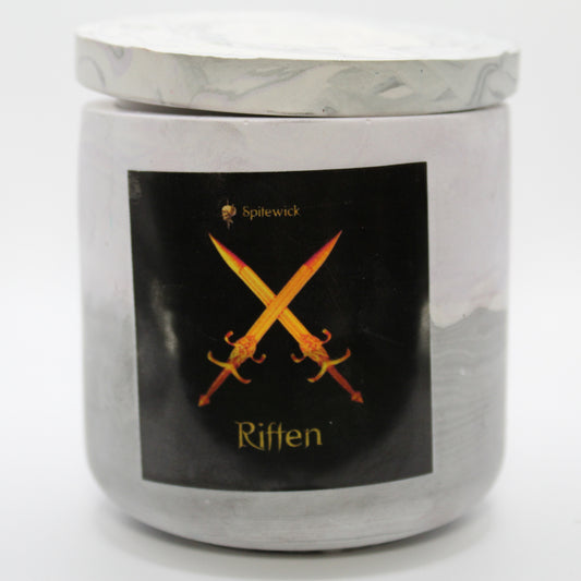 A soft purple and grey candle holder with a sleek black label, featuring two crossed swords intricately designed in stylized gold Celtic knotwork. Beneath the design, the name riften is elegantly printed in gold, adding a bold yet sophisticated touch. Set against a clean white background, this handcrafted candle blends tradition with modern artistry, perfect for unique home decor, gifts, and personal expression.