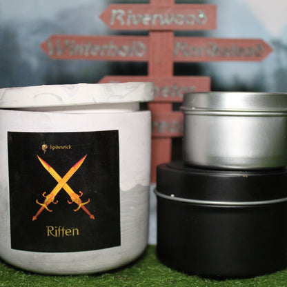 Front view of a handcrafted soft purple and grey candle holder with a black label featuring two crossed swors in gold Celtic knotwork, with the name Riften in gold below. Two smaller candles—one black 4oz and one silver 2oz—show the three available sizes. Set on grass in front of a 3D-printed signpost marking Skyrim cities, blending medieval fantasy with artisan craftsmanship.
