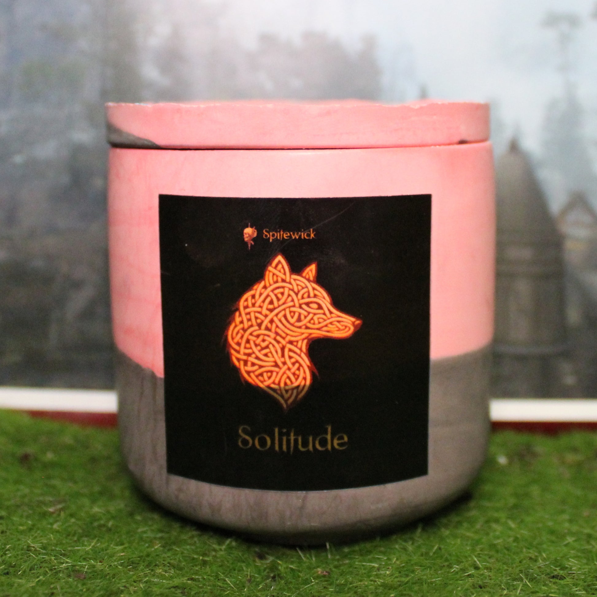 Front view of a handcrafted red and black candle holder with a black label featuring a wolf in gold Celtic knotwork. The name Solitude is elegantly printed in gold beneath the design. Set on grass, this piece blends artisan craftsmanship with bold identity, making it perfect for unique home decor, gifts, and statement lighting.
