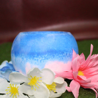Handcrafted candle holder featuring the pink, blue, and white hues of the transgender pride flag, seamlessly blended together for a striking design. Set on grass with a burgundy vinyl backdrop, surrounded by an assortment of flowers mirroring the flag’s colors, creating a visually rich and identity-affirming scene. Perfect for LGBTQ+ home decor, pride gifts, and statement lighting."